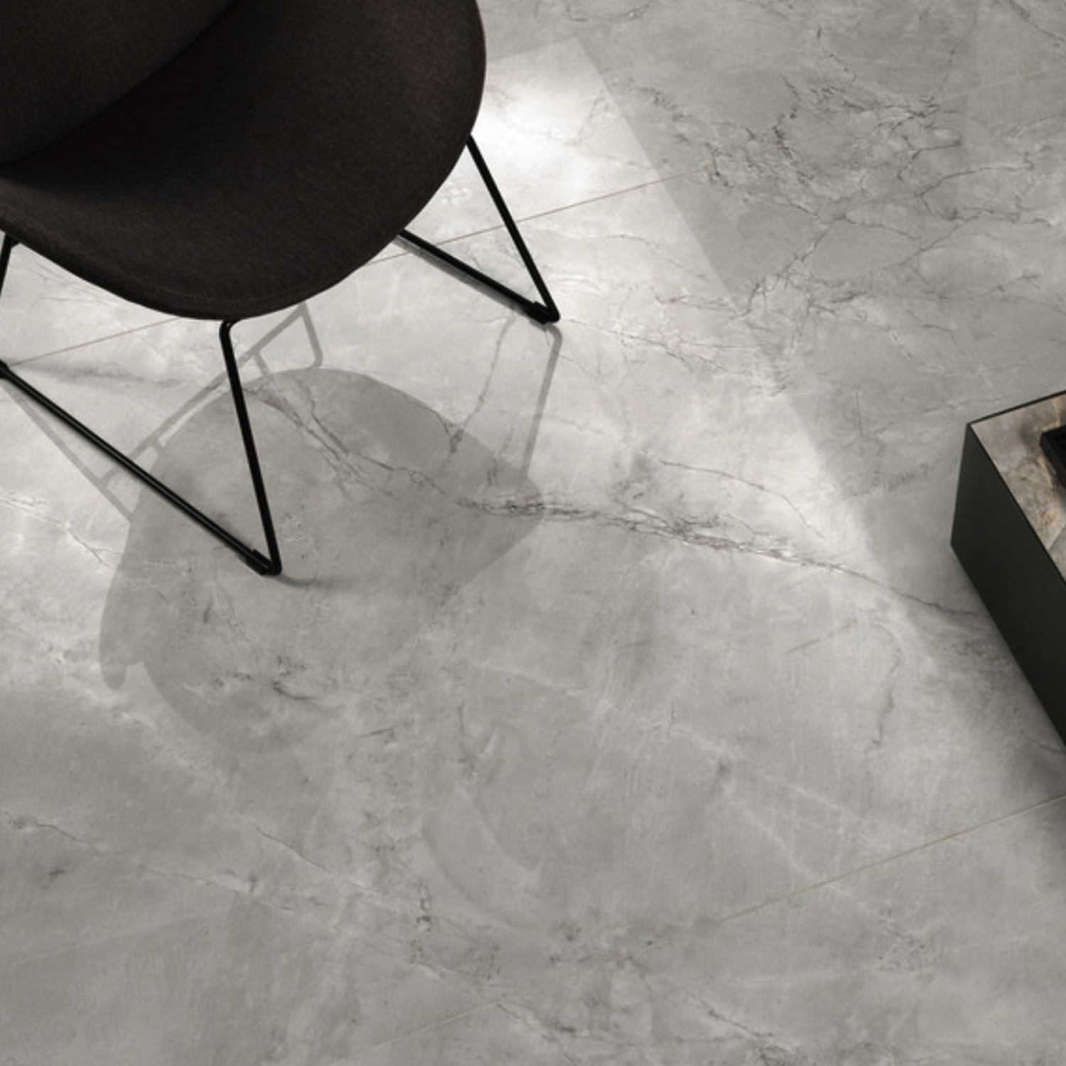 A black chair with slender metal legs rests on a Marvel X - Grey Cloud polished light gray marble floor, featuring natural veining. The edge of a dark rectangular object appears in the lower right corner.