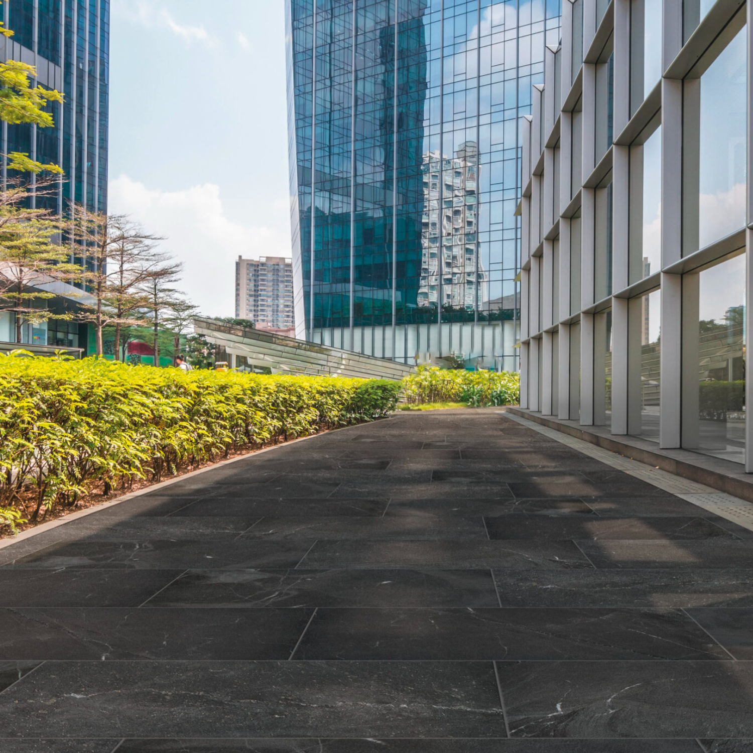 A clean walkway paved with Modern Slate - Slate Charcoal runs alongside green bushes on the left, adjacent to a modern glass building. Towering skyscrapers rise in the background beneath a blue sky dotted with clouds.