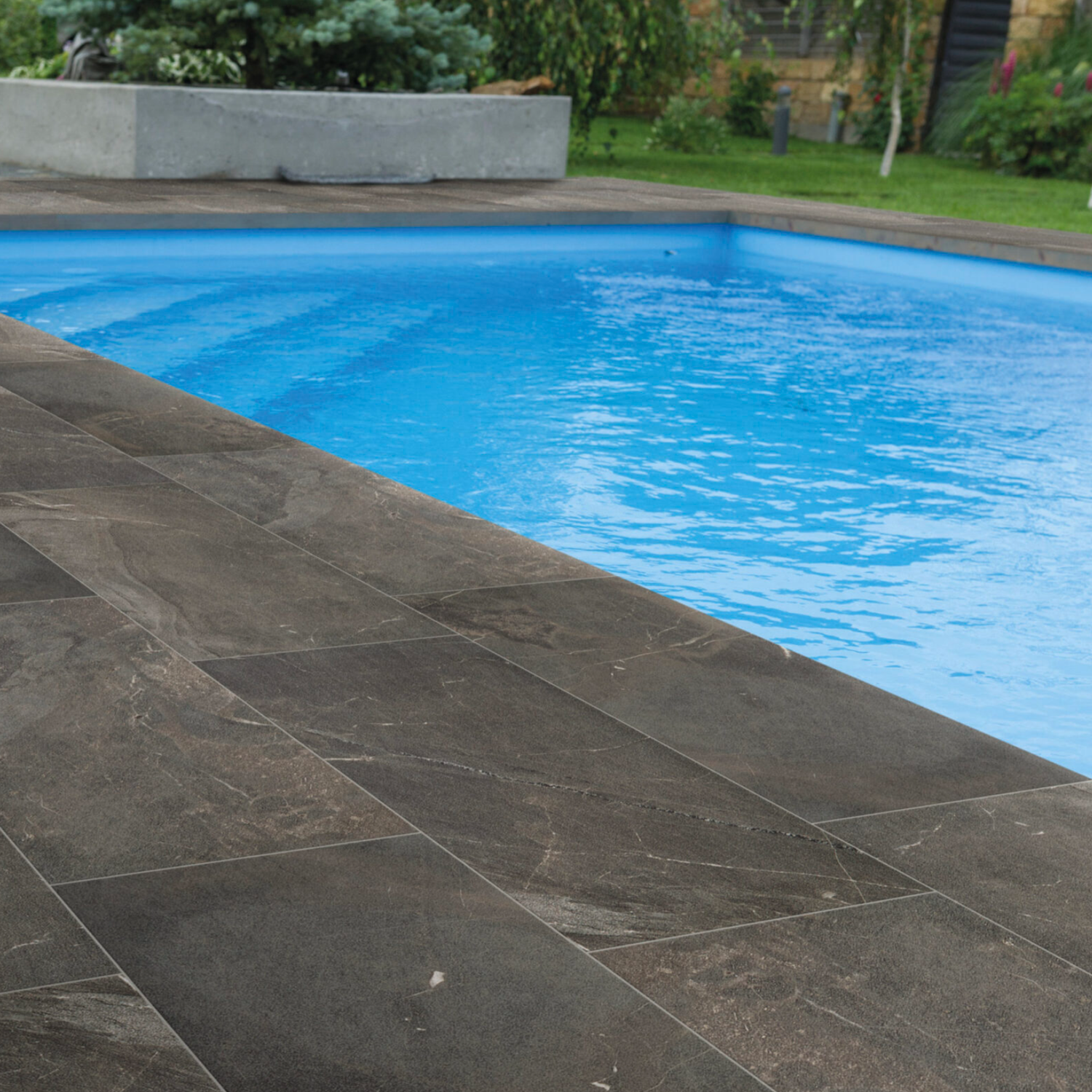A swimming pool with clear blue water is surrounded by large rectangular Modern Slate - Slate Charcoal tiles. Trees and green plants can be seen in the background.