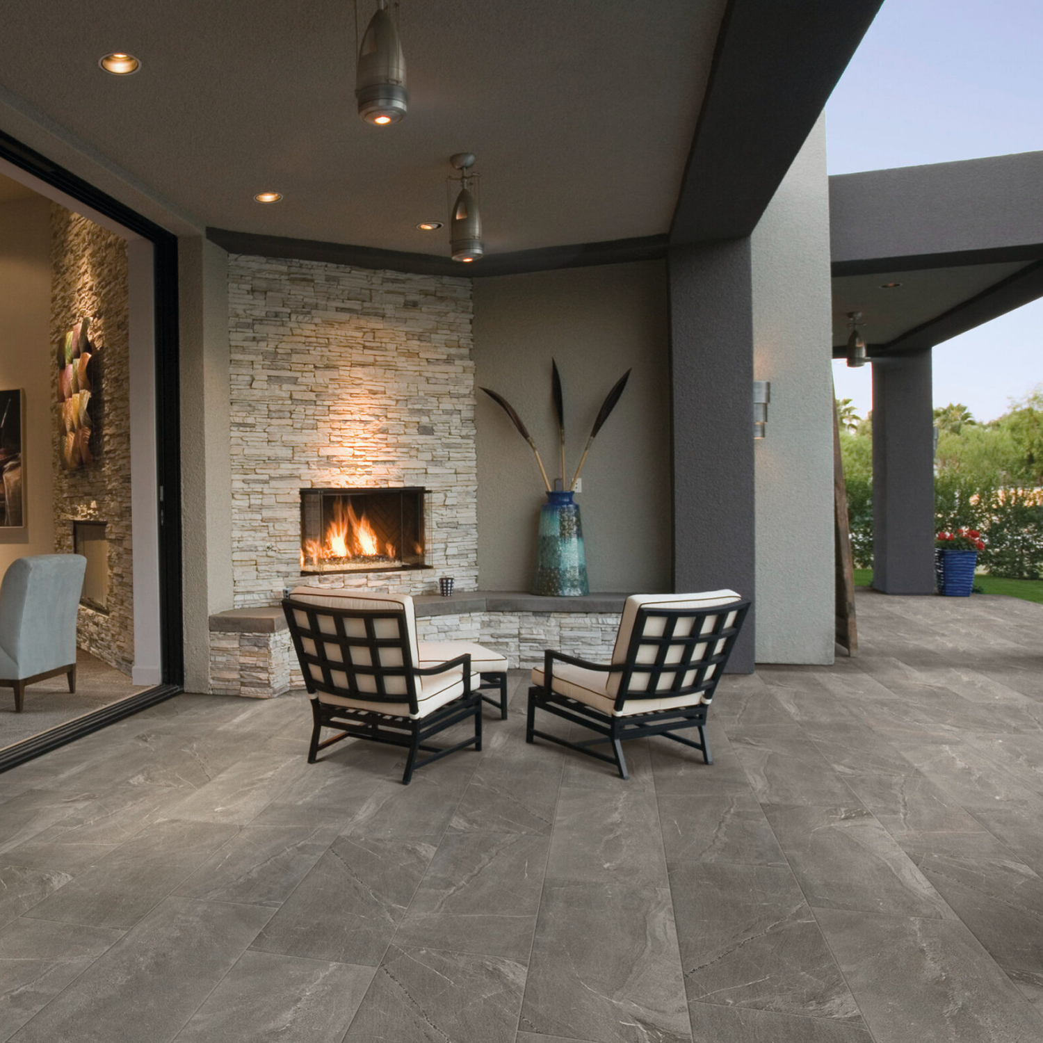 Modern covered patio with stone fireplace, two cushioned chairs, a large blue vase with tall branches, and Modern Slate - Slate Grey tile flooring. Interior room visible through open sliding glass doors. Evening lighting.