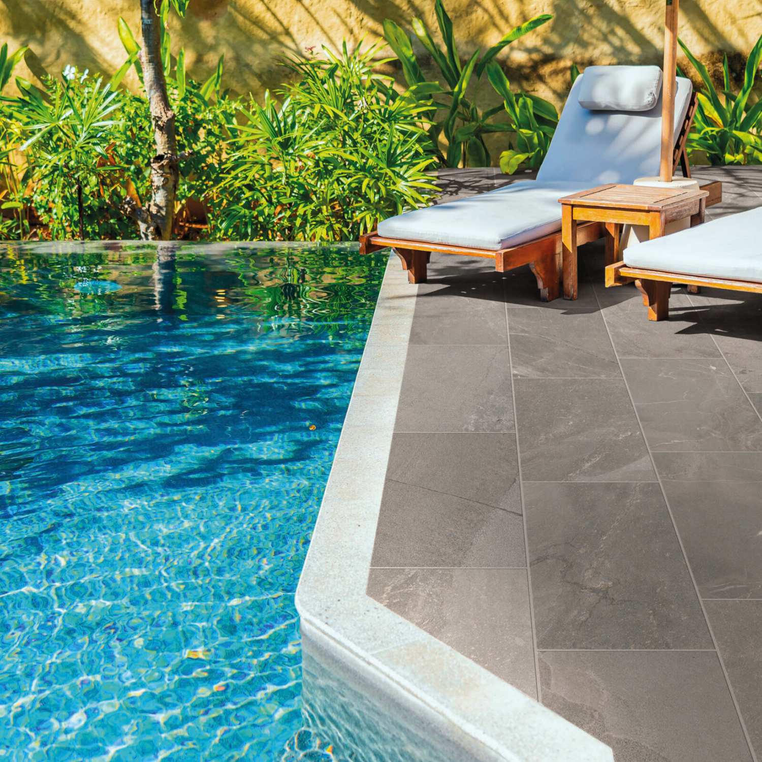 Two Modern Slate - Slate Grey cushioned lounge chairs and a small wooden table sit beside a clear blue swimming pool, surrounded by green plants and sunlight, creating a relaxing outdoor scene.