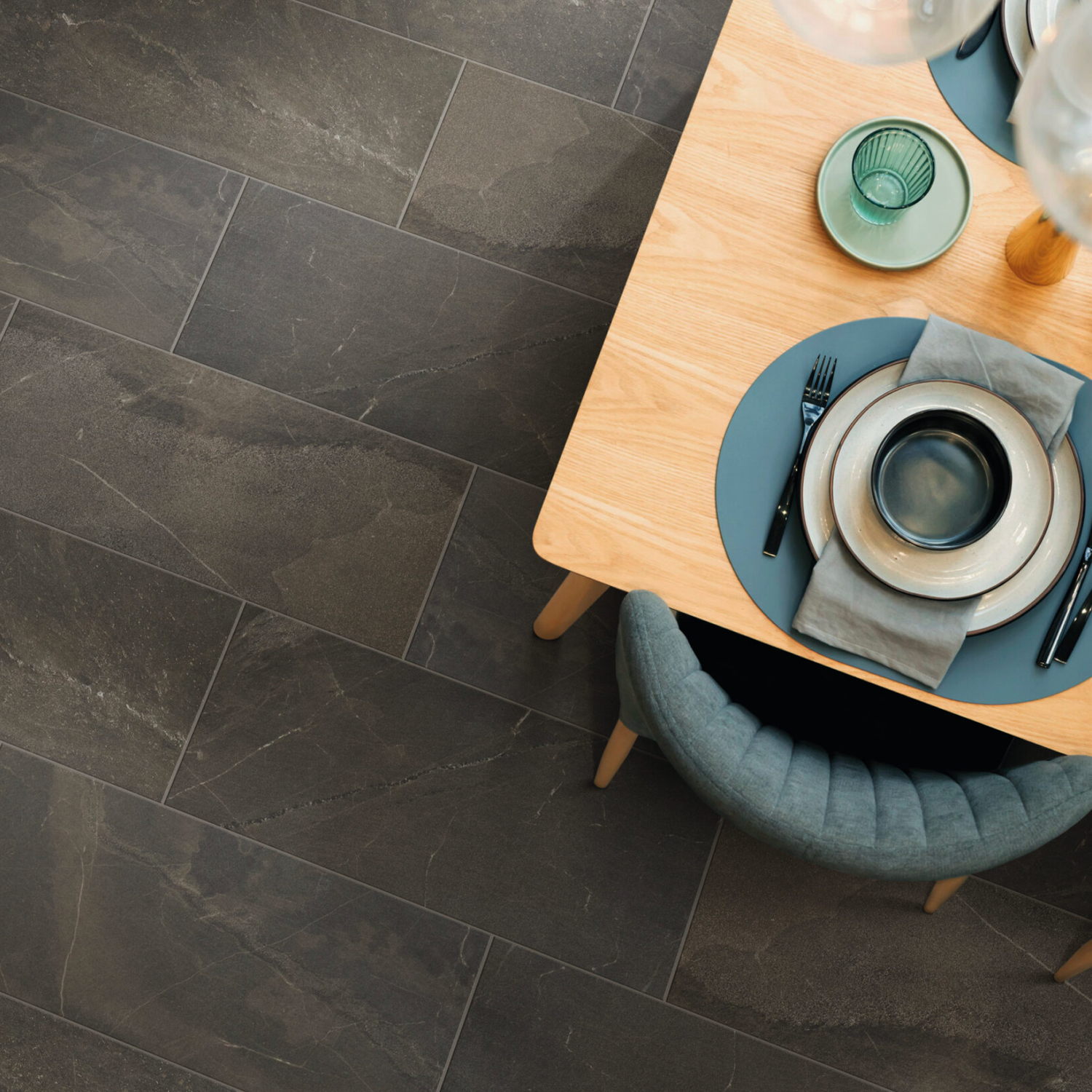 A wooden dining table set with plates, cutlery, and glasses on gray placemats, beside a cushioned chair, sits on a floor featuring Modern Slate - Slate Noce tiles.