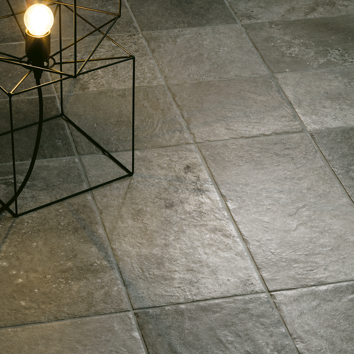 The Molo Audace - Bocca Di Lupo, an illuminated exposed bulb lamp with a wireframe base, rests on a gray stone tile floor, casting gentle light and shadows across the textured surface.