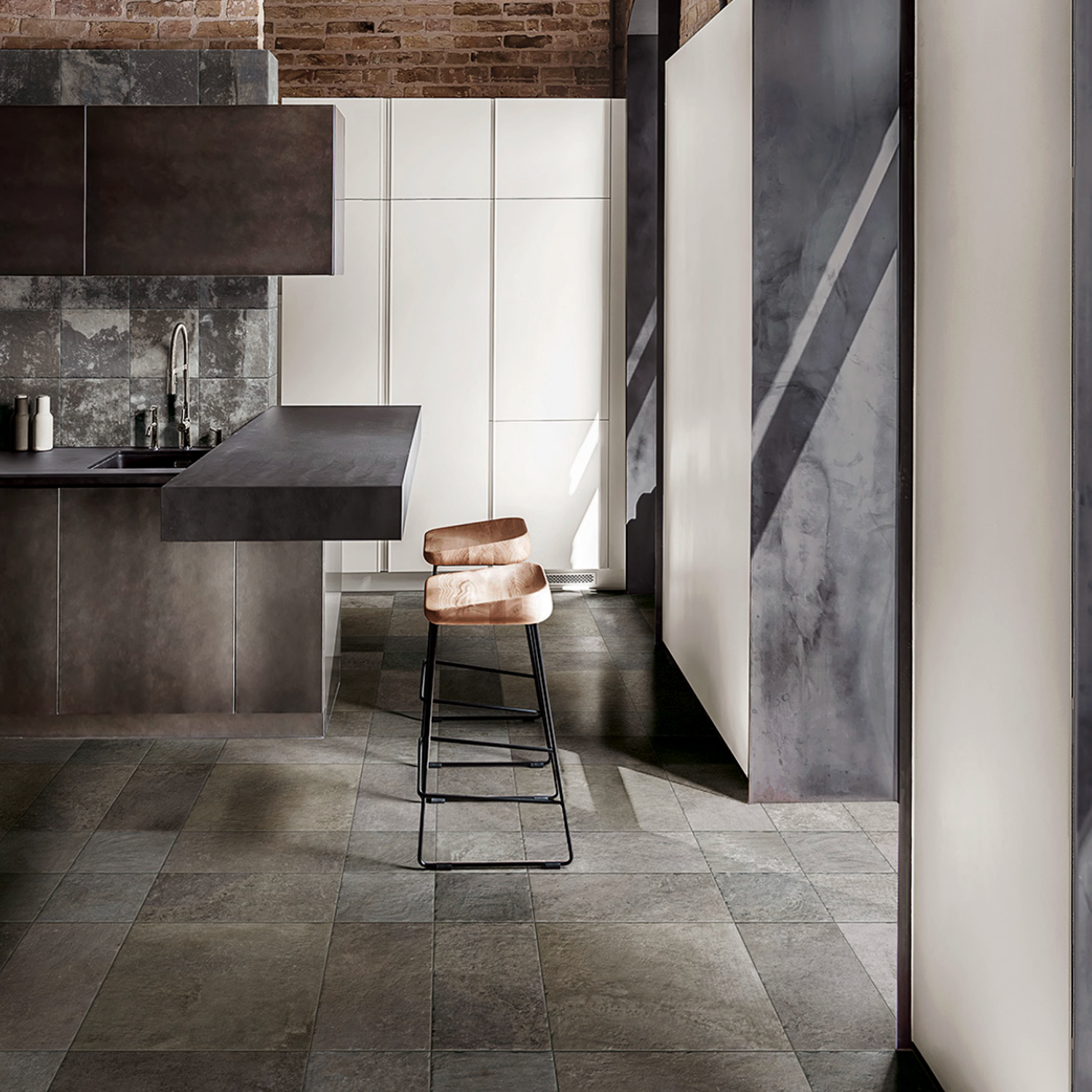 A modern kitchen featuring dark cabinets and a Molo Audace - Bocca Di Lupo stone backsplash, complemented by minimalist decor, two cushioned barstools at the counter, large floor tiles, and sunlight casting shadows on crisp white walls.