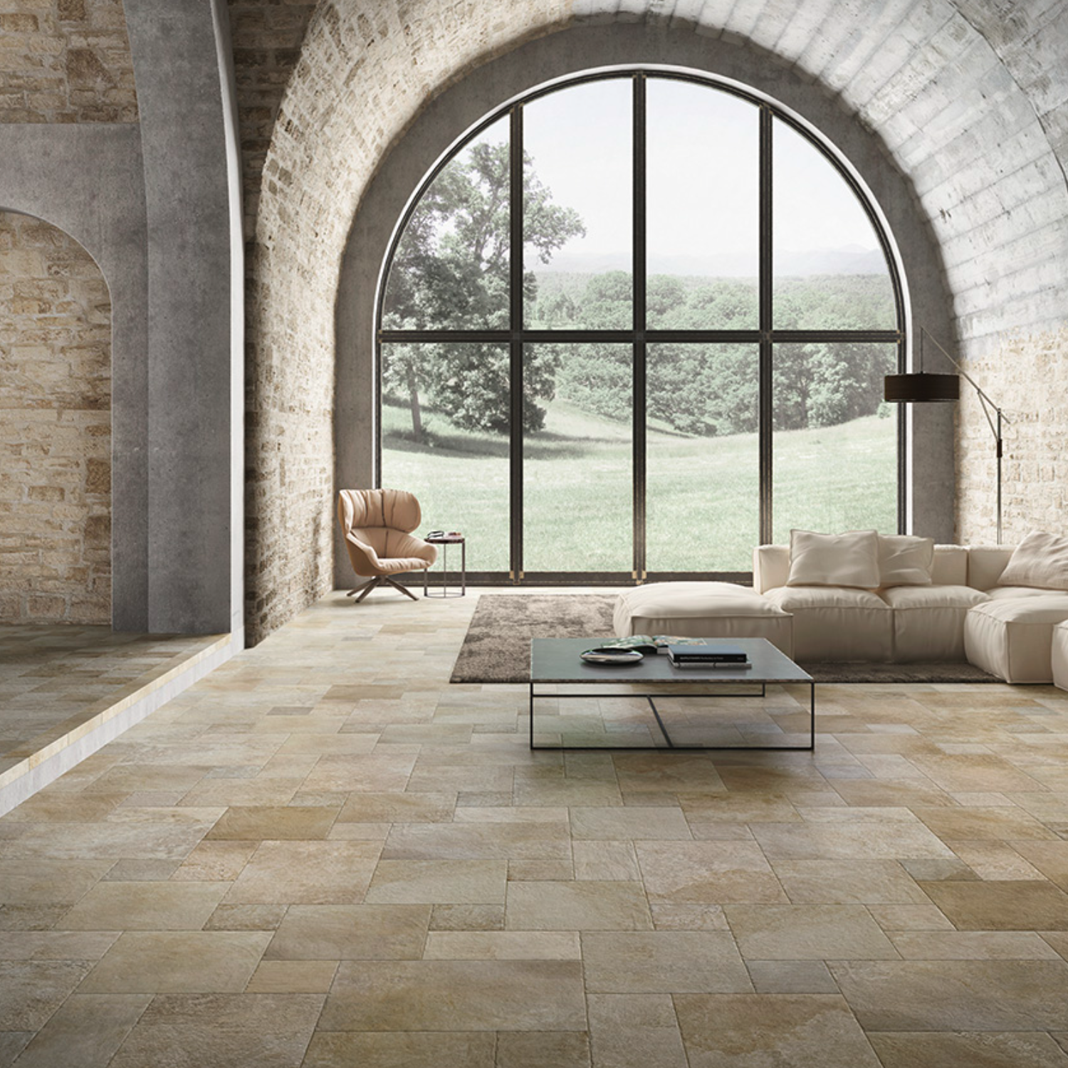 Transformed sentence using product data: Spacious living room with stone walls and arched window features a modern beige sofa, armchair, and coffee table beautifully set on a Molo Audace - Terra D'Ormeggio tiled floor. A large window frames views of the lush green landscape outside.