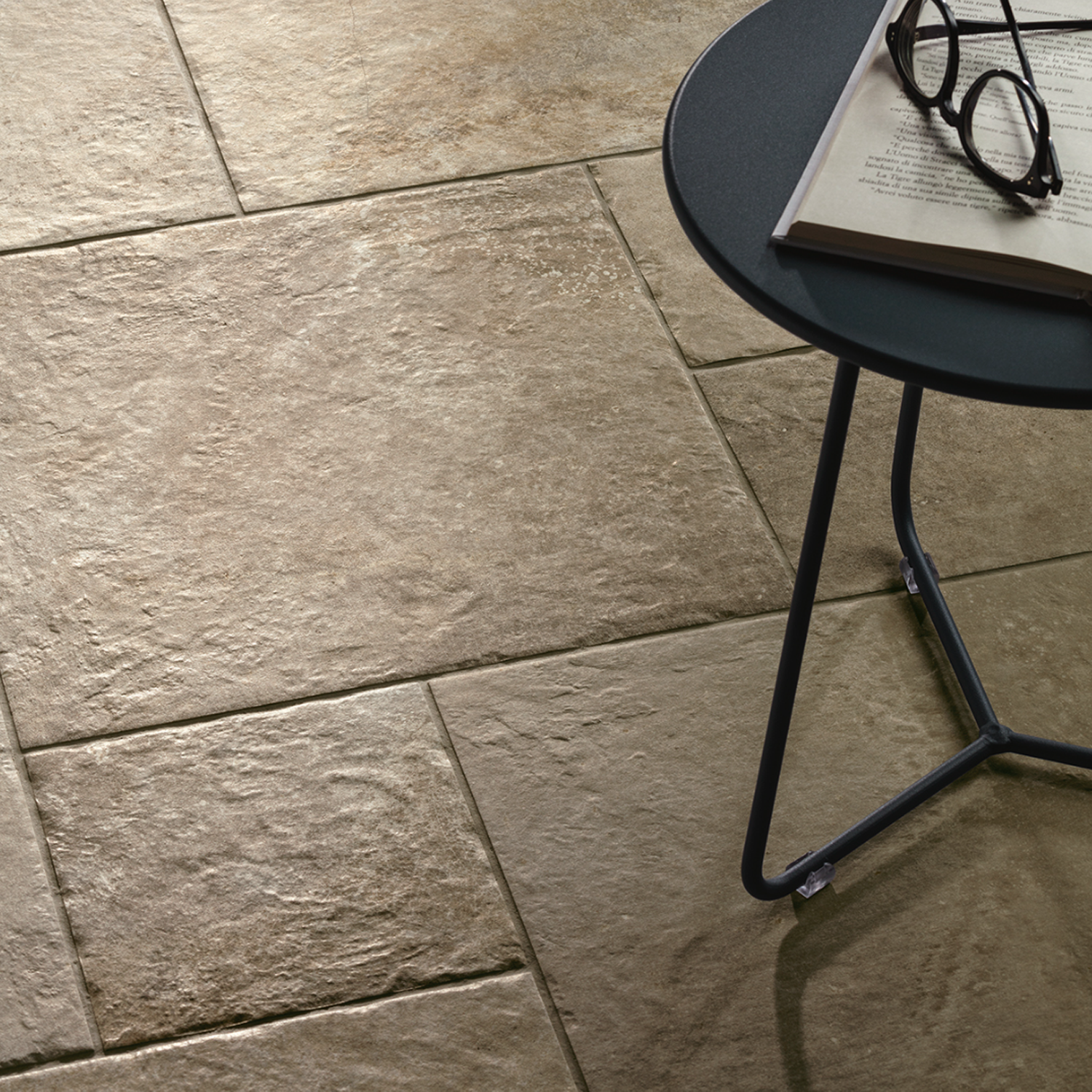 A round black table with an open book and eyeglasses sits on a floor of large, textured Molo Audace - Terra D'Ormeggio tiles.