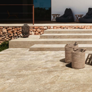 Modern outdoor patio featuring Norde - Oro beige stone tiles, woven baskets, steps leading to a raised seating area with black lounge chairs, a small wooden table, and a stone wall with greenery on the left.
