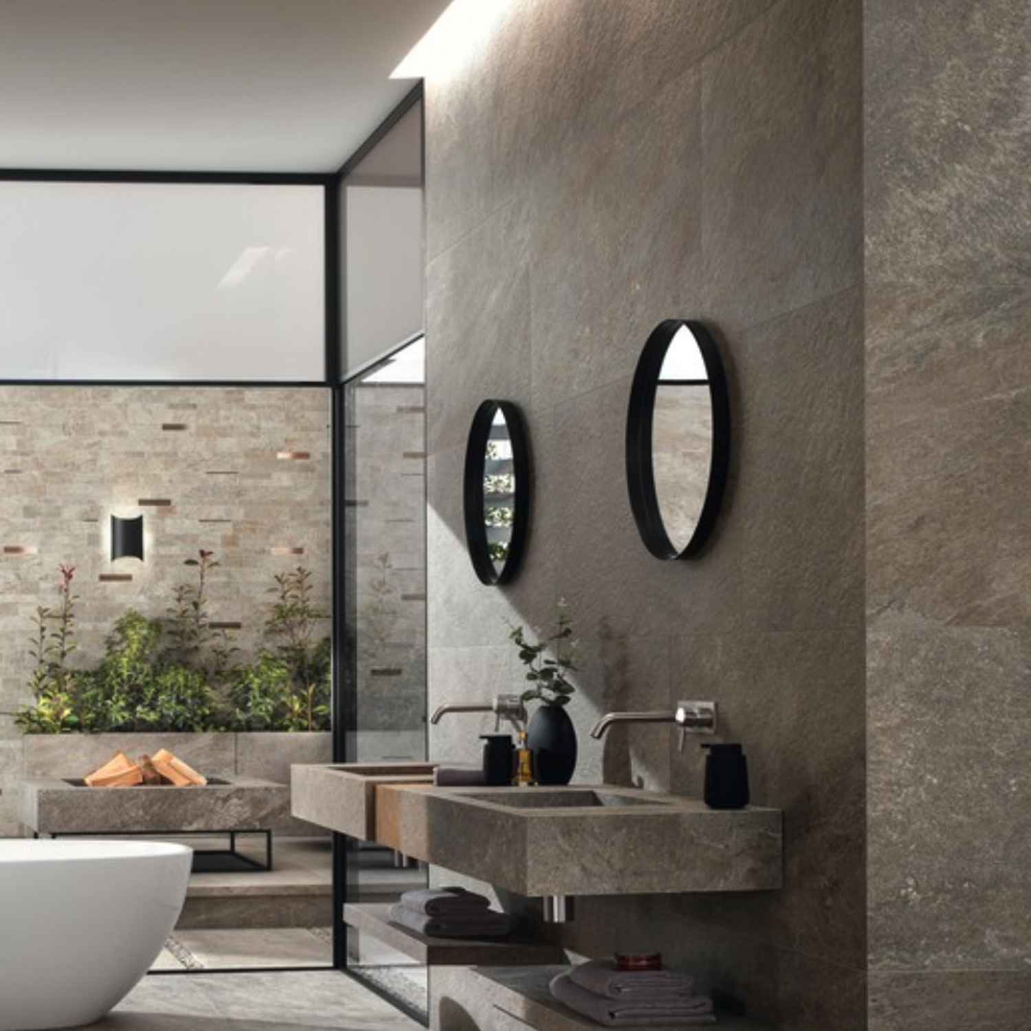 Modern bathroom featuring stone-look Norde - Piombo walls, two oval mirrors, double sinks, a white bathtub, and large windows that open to a walled outdoor garden with greenery and stacked firewood.