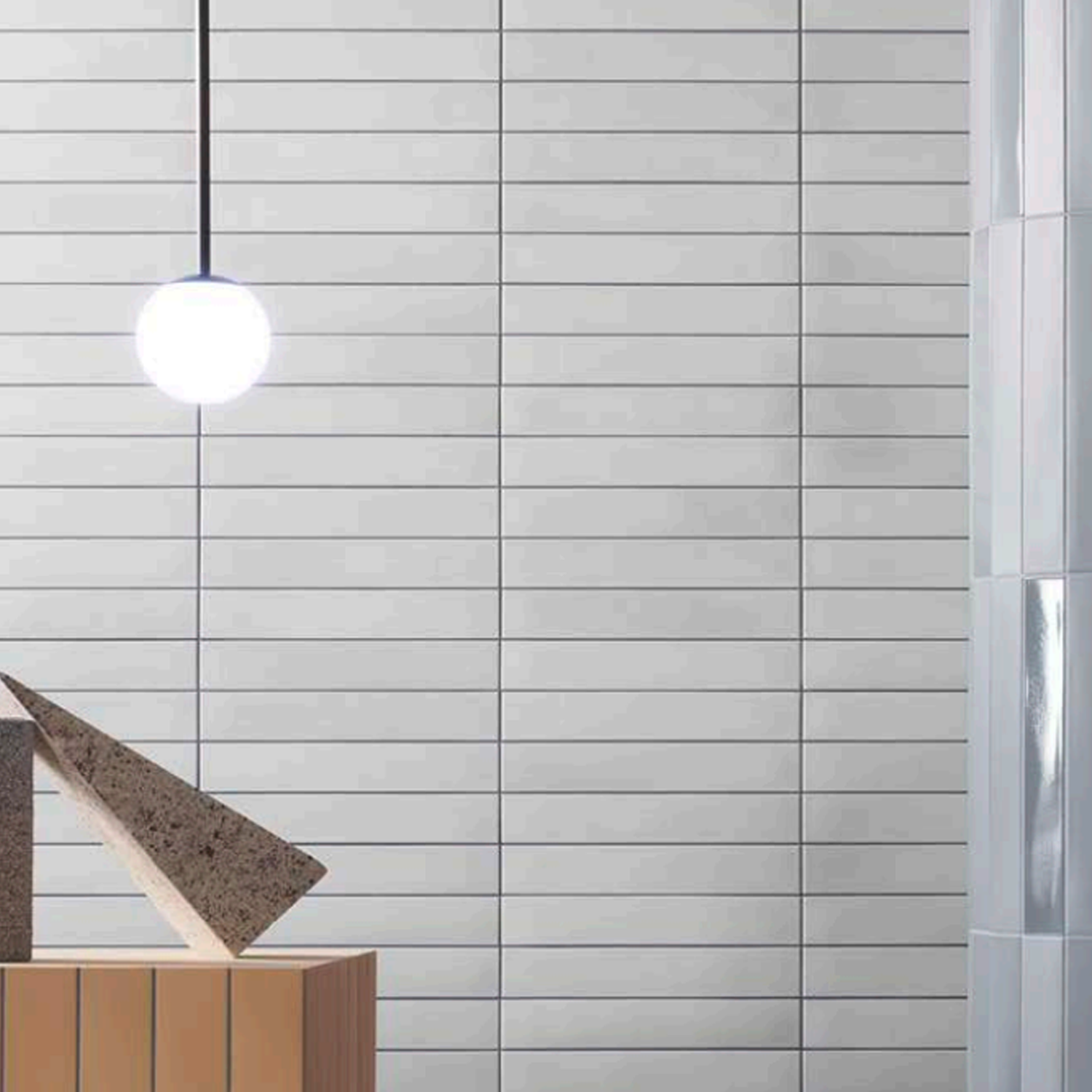 A modern minimalist interior featuring the Pigmento - Bianco pendant light glowing above, accompanied by geometric wooden and stone shapes, a tiled wall, and a reflective column on the right.