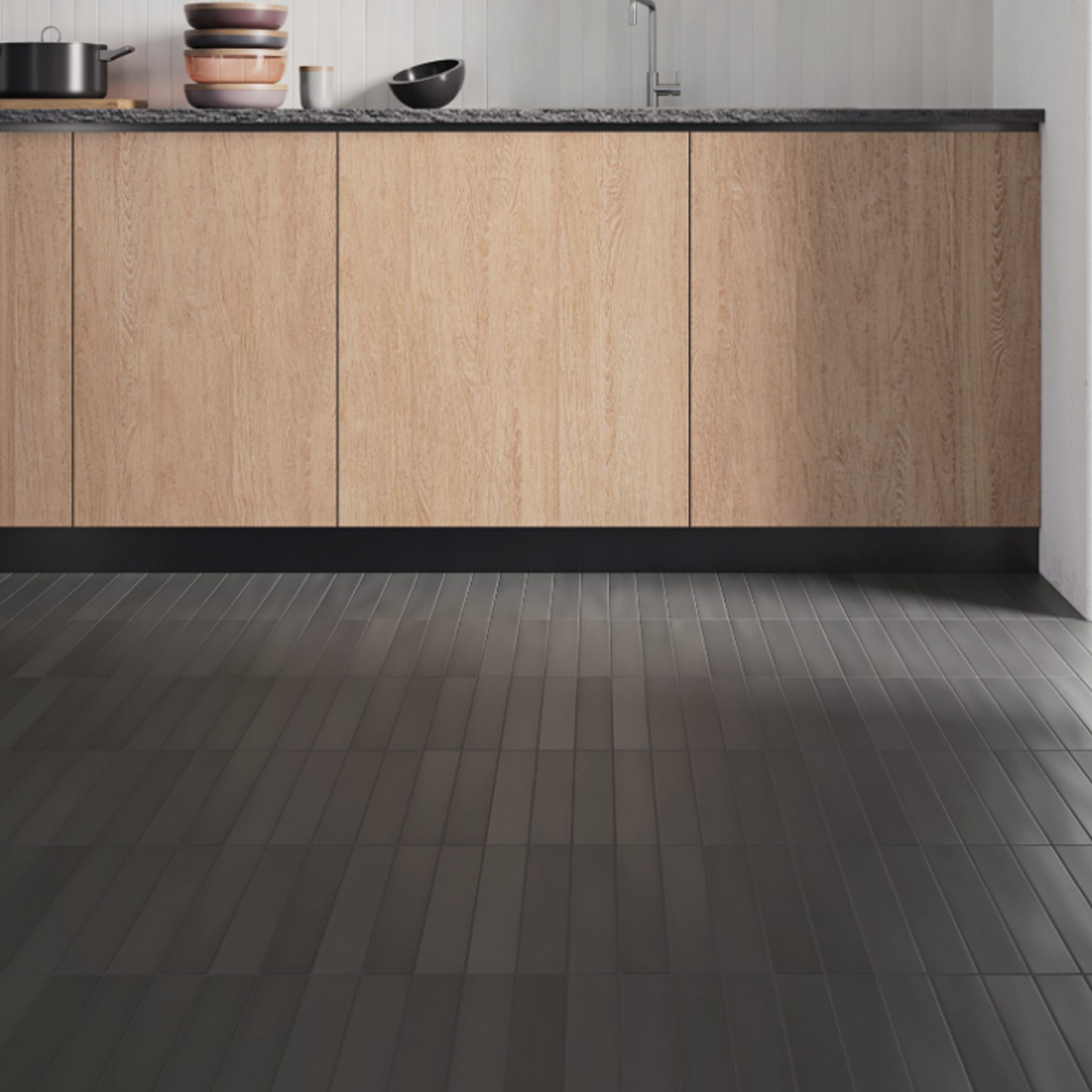 Modern kitchen with light wood cabinets, black countertop, and a black Pigmento - Fango tile floor. A stack of pots and a bowl sit on the counter beside a stainless steel sink and faucet. White wall panels are in the background.