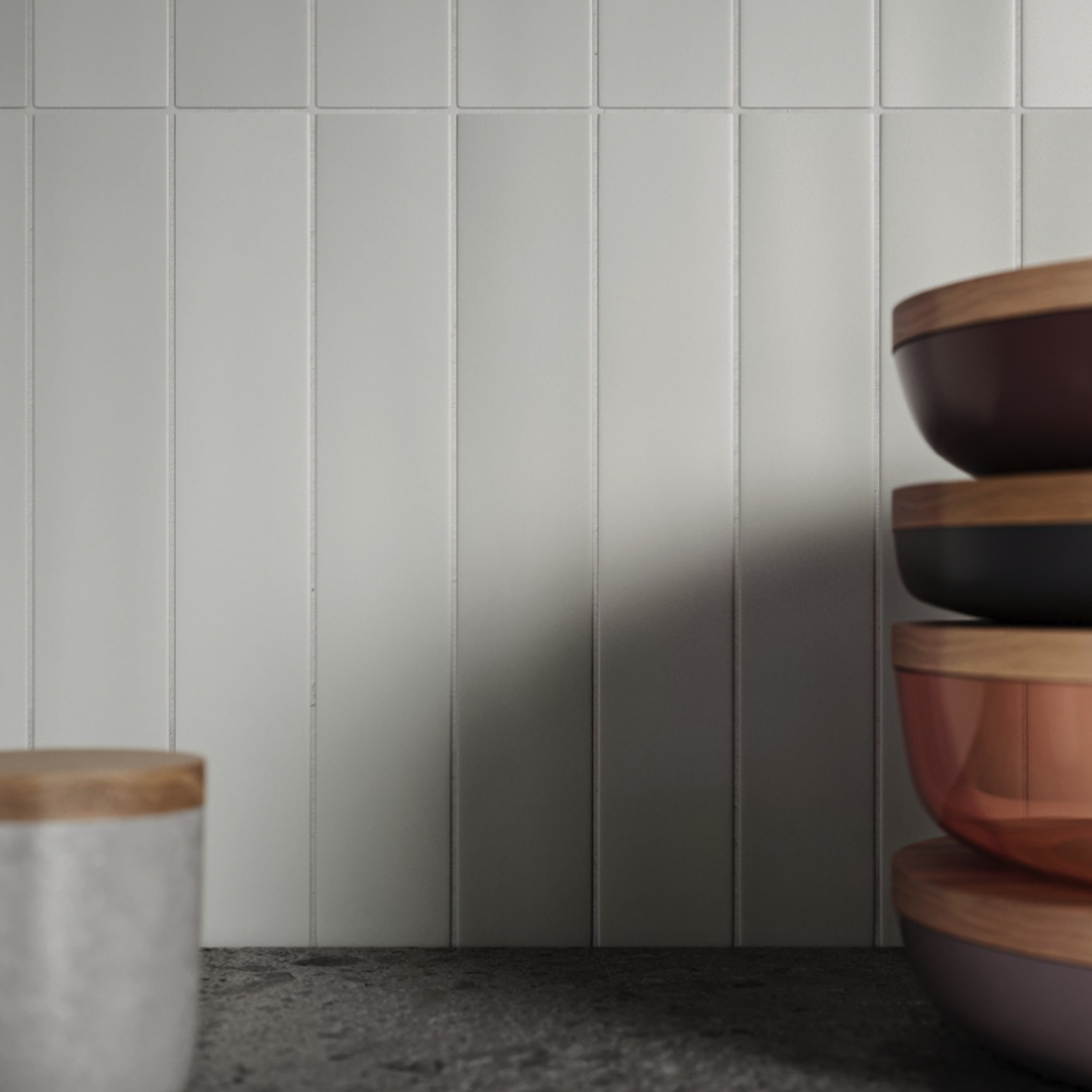 Four stacked Pigmento - Grigio bowls with wooden lids are on the right side of a gray textured countertop, next to a white cup with a wooden lid. The background features white vertical tile.