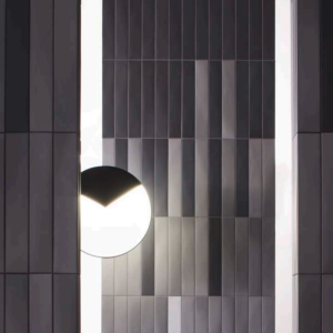 A round mirror with a shadowed edge is mounted on a wall finished in Pigmento - Nero, featuring vertical panels in shades of gray that create a geometric, modern architectural look.