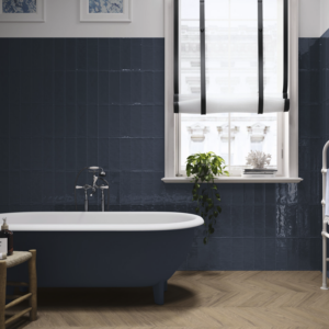 A bathroom featuring the Pim Pam Pum - Pum Blue clawfoot tub, complemented by dark blue wall tiles, a window dressed with a black-and-white shade, and potted plants arranged on the windowsill.