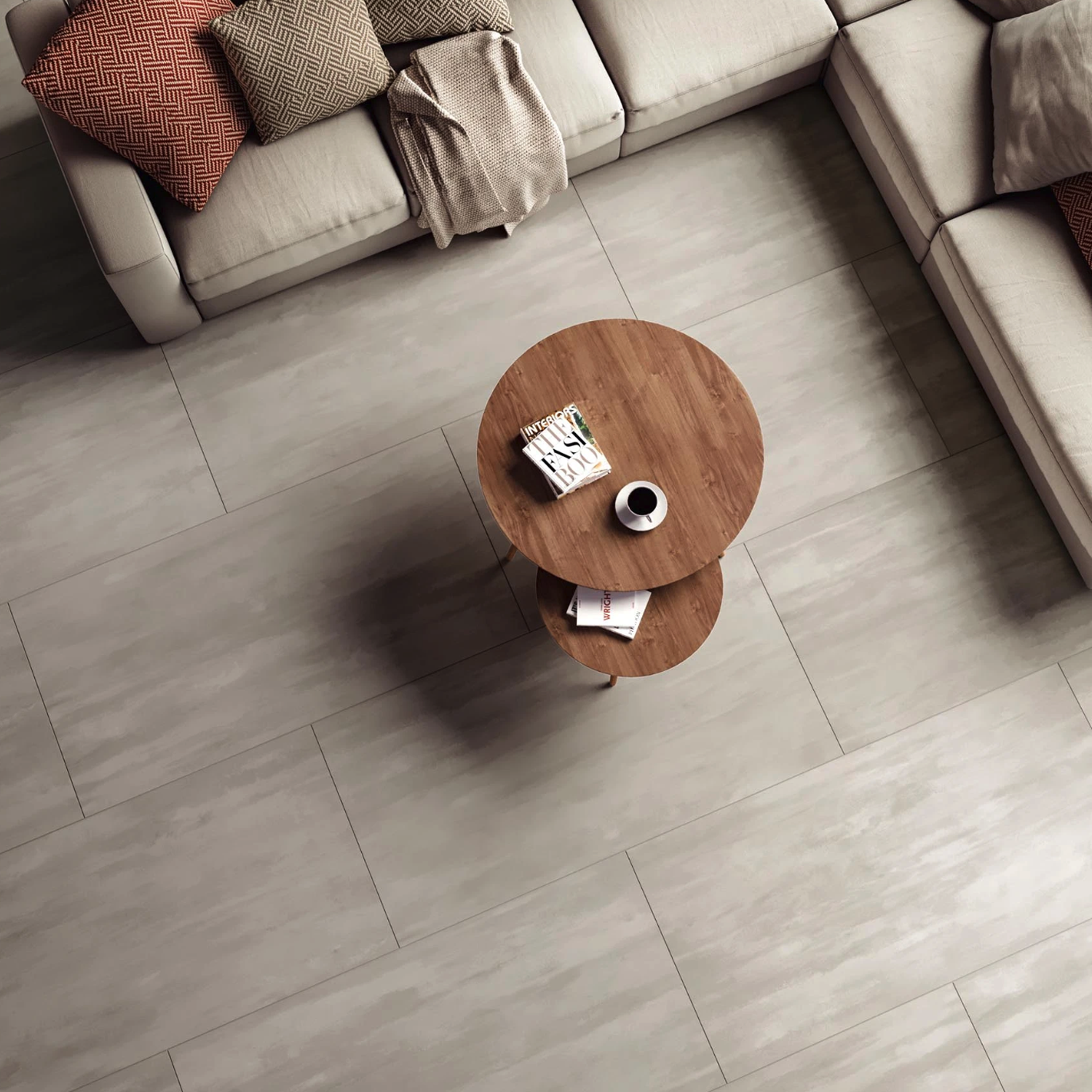 A top-down view of two round Reflex - Mercury wooden tables with a cup, book, and magazine, surrounded by a beige sectional sofa adorned with red and patterned cushions on a light gray tiled floor.