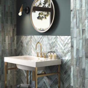 Modern bathroom featuring Savannah - Berilo greenish-blue herringbone and vertical tile walls, round mirror, gold faucet, white sink, glass jars, wall-mounted light fixture, and a window reflecting tree branches.