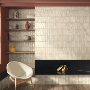 A modern living room featuring a Seville - Bone tiled wall, a fireplace with stacked firewood, four wooden shelves displaying books, pottery, and stones, and a cream armchair on a wood floor. The surrounding walls are painted in warm terracotta tones.