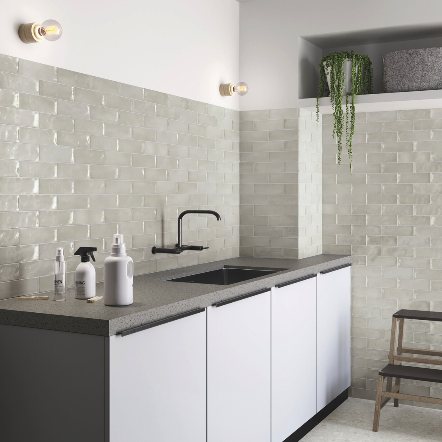 Modern kitchen featuring a Seville - Grey subway tile backsplash, black faucet, soap dispensers, and white cabinets. A potted plant sits on a high shelf, with a small wooden stool beside the counter. Two wall lights are visible.