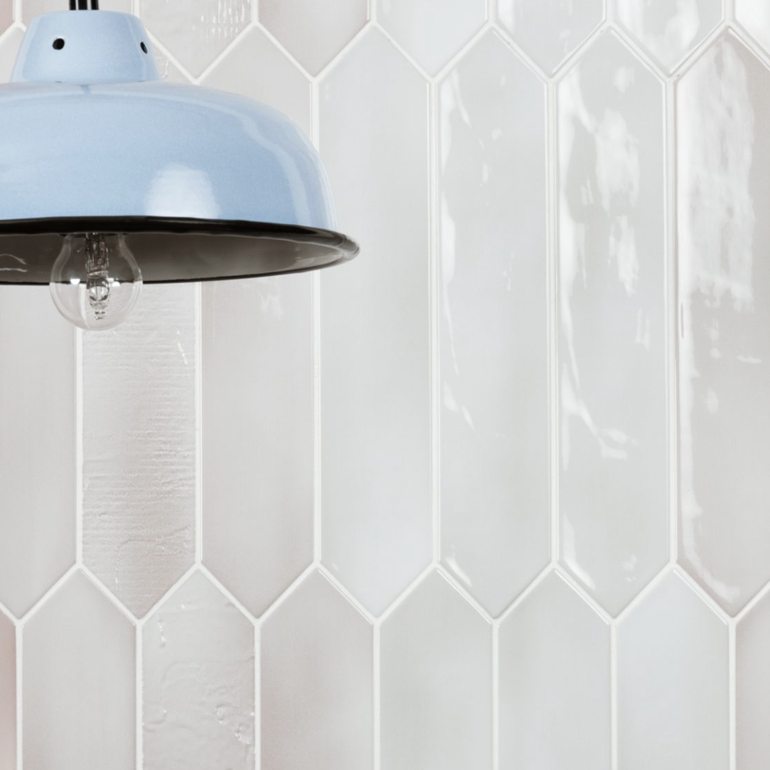 A Sfumature - Grigio pendant light in pale blue hangs in front of a white, glossy, elongated hexagon tile wall, with only part of the lamp visible in the upper left corner of the image.