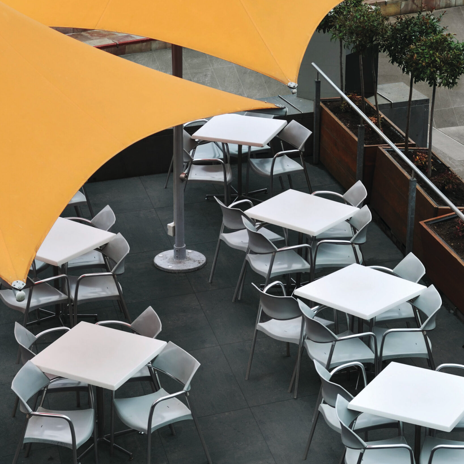 The Simply Modern - Black outdoor patio features several empty white tables and chairs arranged beneath large yellow shade sails, set next to rectangular planters with small trees on a tiled floor, creating a clean and modern atmosphere.