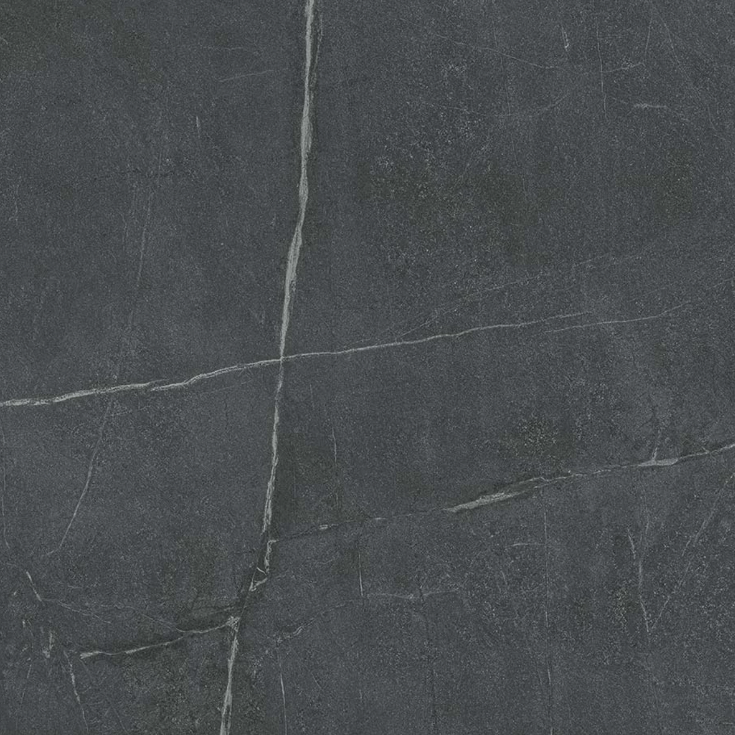 Soapstone Dark features a dark gray stone surface with thin, light-colored natural veins and subtle texture, closely resembling slate or marble. Its pattern includes intersecting lines and a matte, slightly rough finish, capturing the authentic look of natural stone tile.