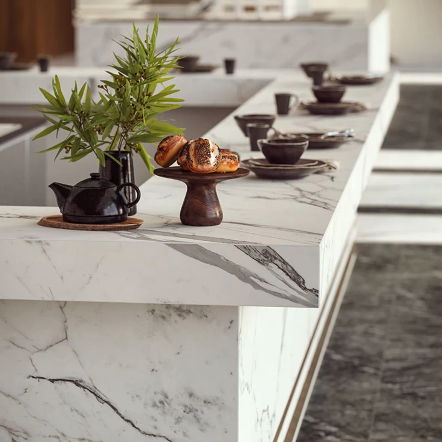 A modern kitchen counter set featuring Statuario Supremo marble, complemented by black dishware, a teapot, a wooden cake stand with pastries, and a small vase with green leaves. The floor also showcases a dark marble pattern.