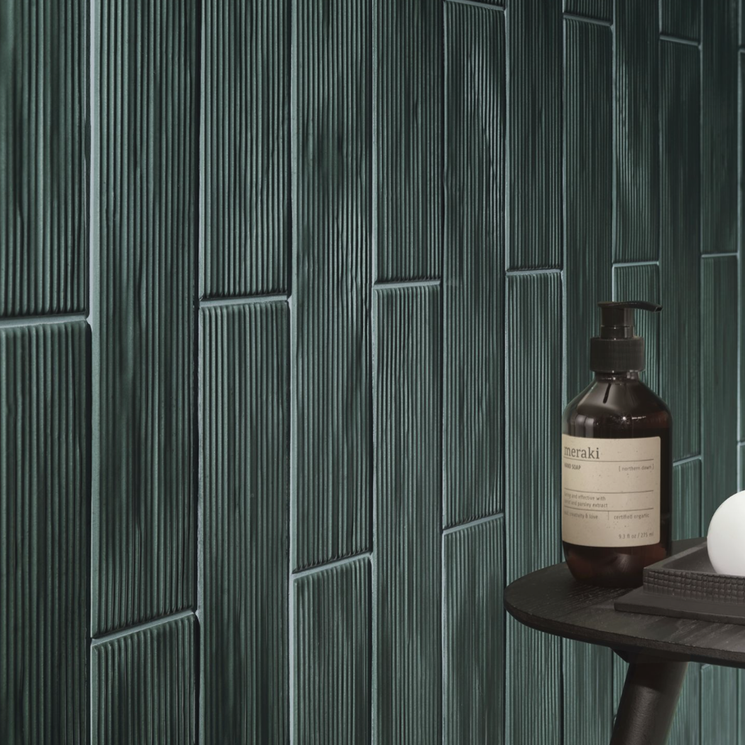 Dark green Swing - Musk vertical tiles with subtle grooves cover a wall. In the foreground, a black shelf holds a brown bottle with a label, a white round object, and a dark rectangular item, suggesting a bathroom or spa setting.