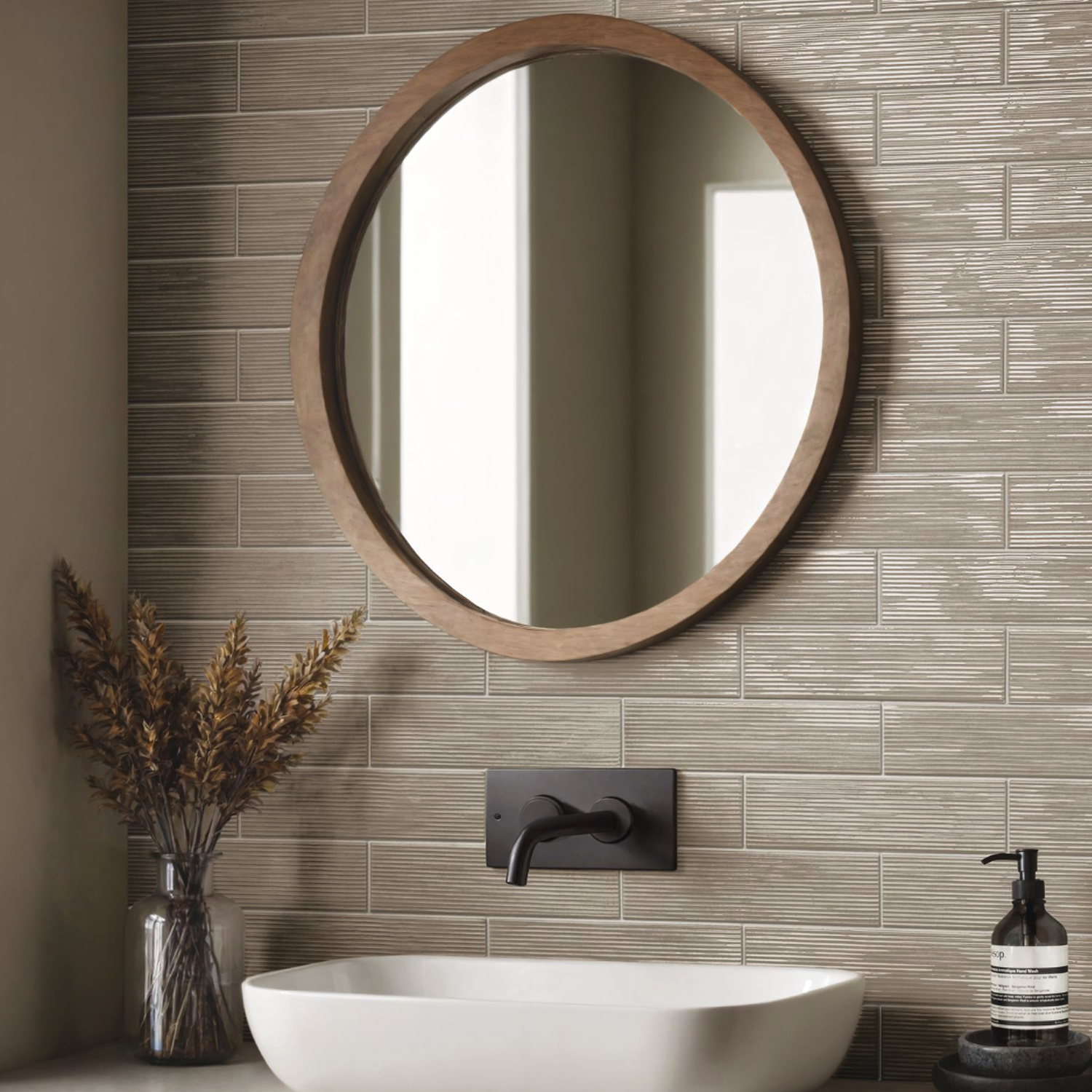 A round Swing - Sand wooden-framed mirror hangs above a white rectangular sink with a black faucet, set against a brown tiled wall; on the counter, there is a soap dispenser and dried plants in a vase.