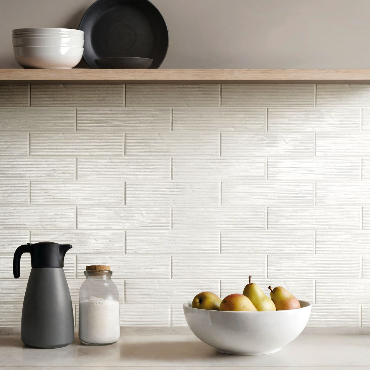 A kitchen counter is adorned with a bowl of pears, a black carafe, and a jar of flour. Above, a wooden shelf displays neatly stacked bowls and plates. The backsplash features Swing - White rectangular, textured tiles.