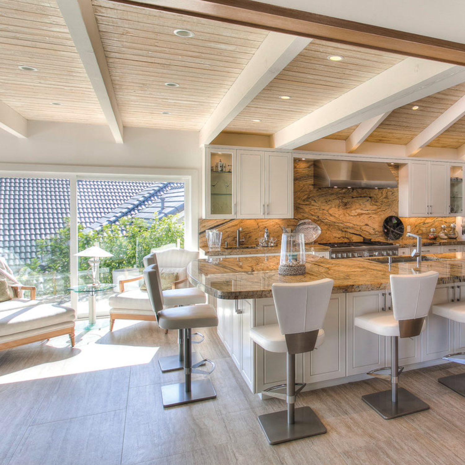 A modern kitchen featuring Travertino Al Contro - Argento Al Contro countertops, sleek white cabinets, three cushioned bar stools, and expansive windows that fill the space with natural light. The cozy seating area overlooks lush greenery outside.