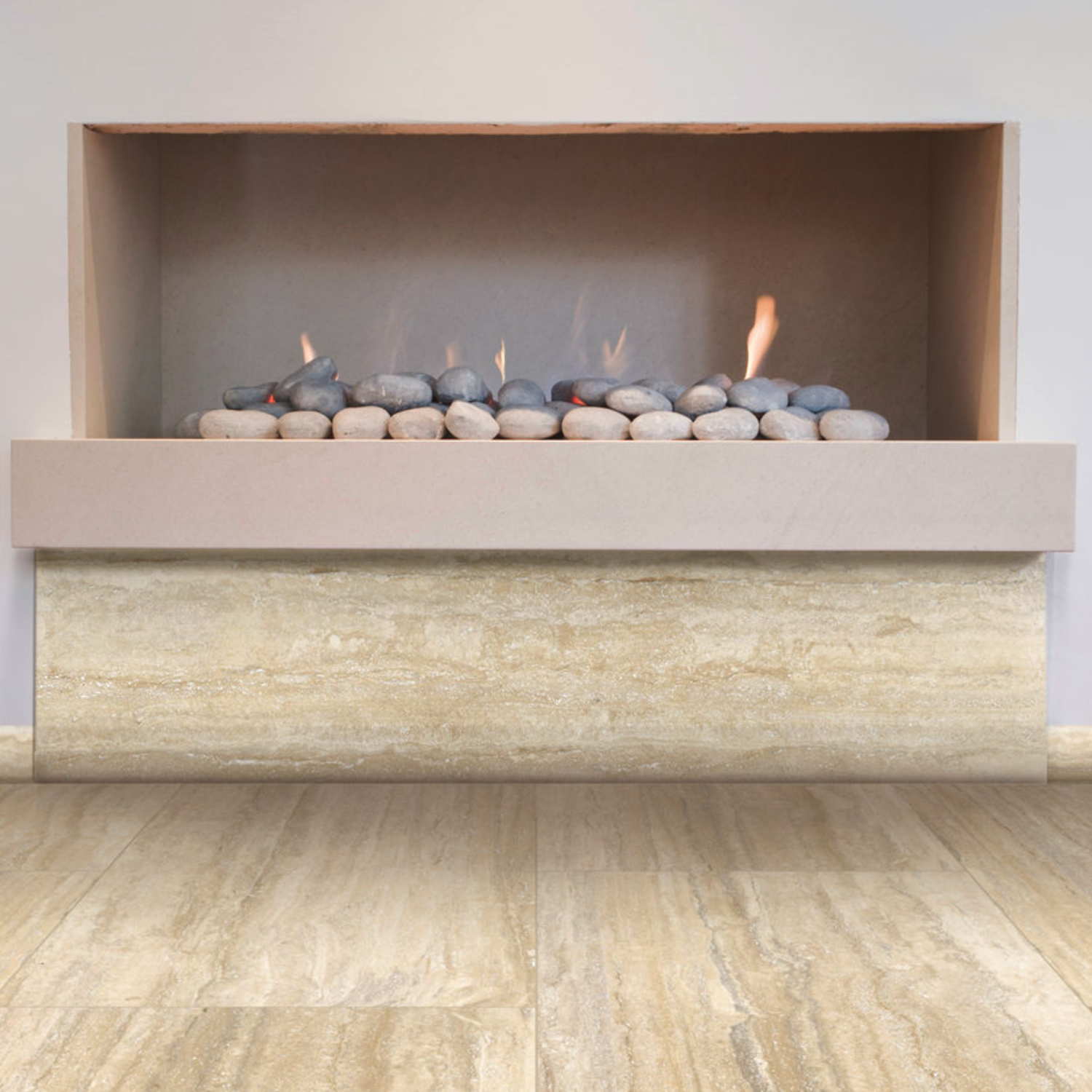 A contemporary gas fireplace features Travertino Al Contro - Argento Al Contro stone with a smooth beige finish, complemented by gray pebbles and gentle flames. Both the hearth and floor are crafted from matching light-colored Argento Al Contro stone tiles.