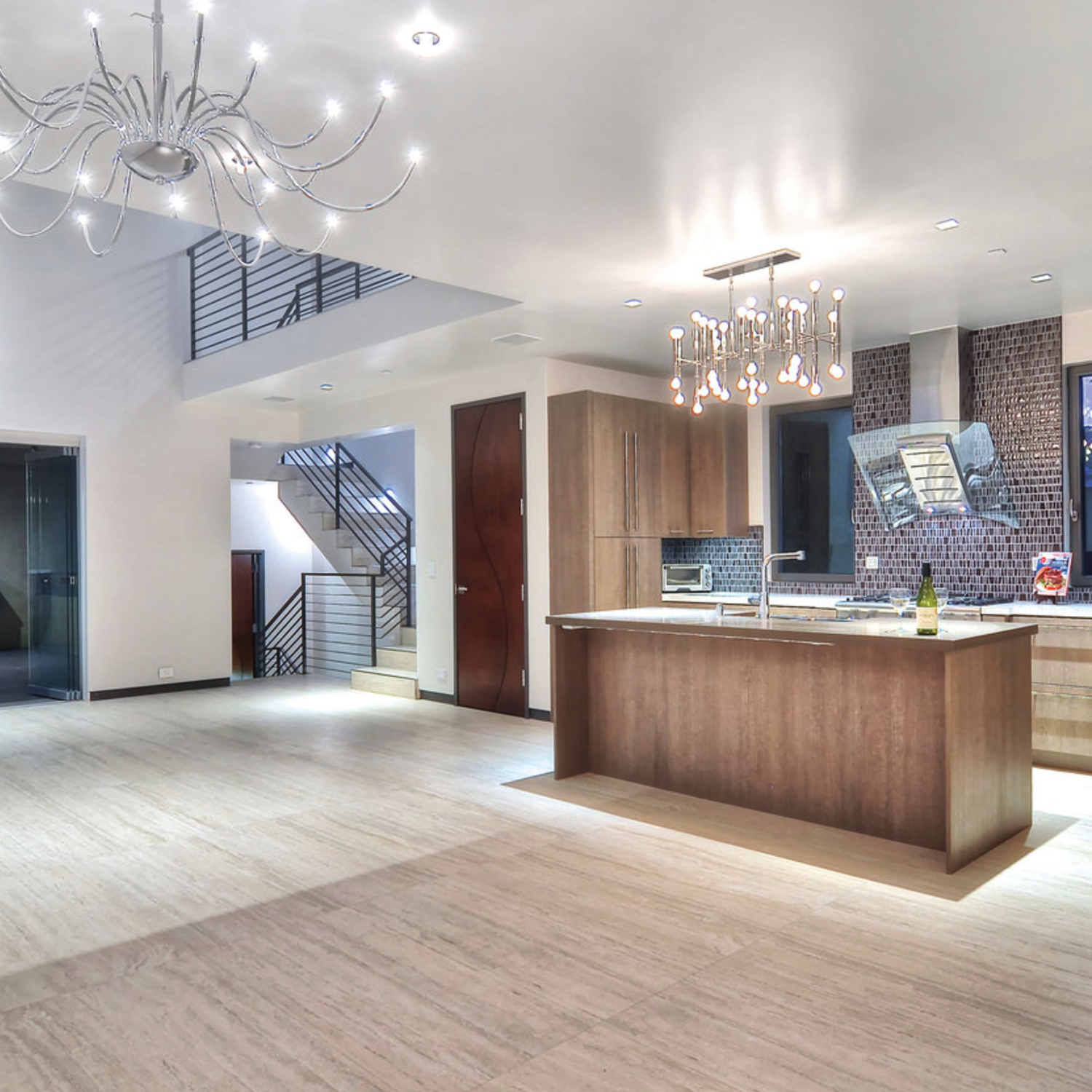 Modern open-concept kitchen with wood cabinets, a central island topped with a bottle of wine and glasses, and hanging chandeliers. The space features Bianco Al Contro tiled flooring by Travertino Al Contro throughout, with a staircase featuring metal railing in the background.