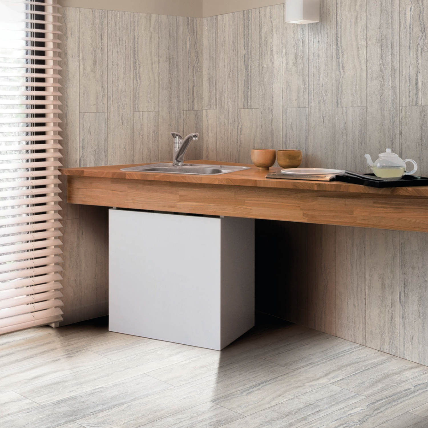 A modern kitchen with light wood floors, a wood countertop, a sink, wooden bowls, and a teapot with cup on a tray. Vertical blinds cover the window on the left, while the walls are finished with Travertino Al Contro - Grigio Al Contro tiles in a neutral tone.
