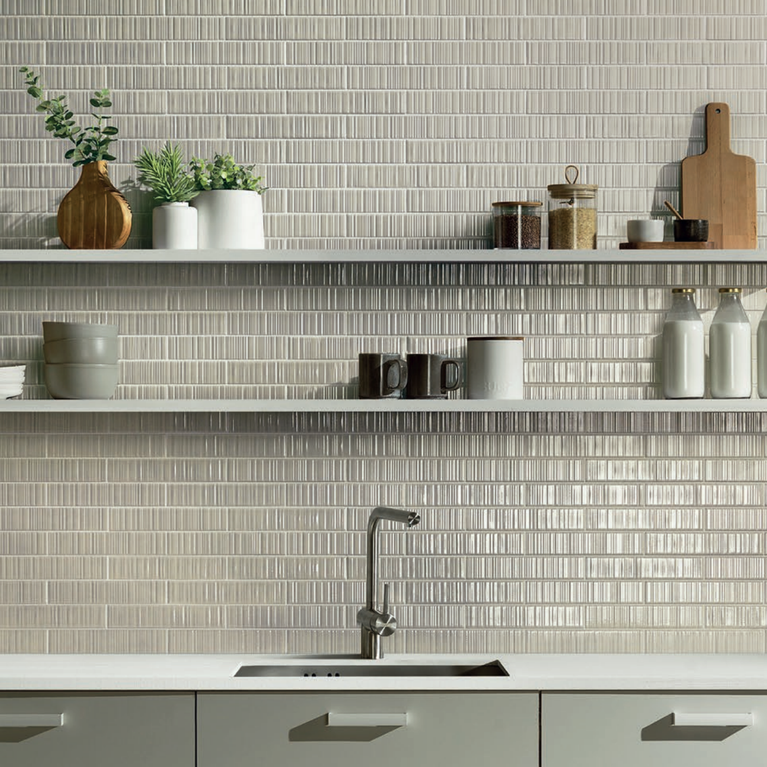 Minimalist kitchen featuring a Vibrant - Beige light gray tile backsplash, open shelves adorned with plants, jars, bottles, and cutting boards. A white countertop with a stainless steel sink and silver faucet sits just below the shelving.