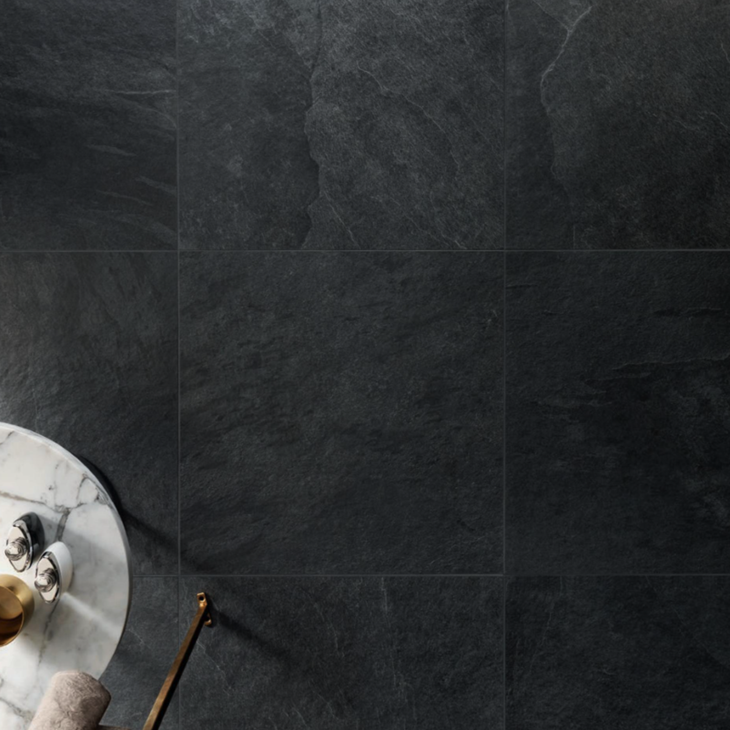 Waterfall - Dark Flow tiles cover most of the floor, with a round marble side table in the lower left corner holding a towel, a gold tray, and a few small bottles.