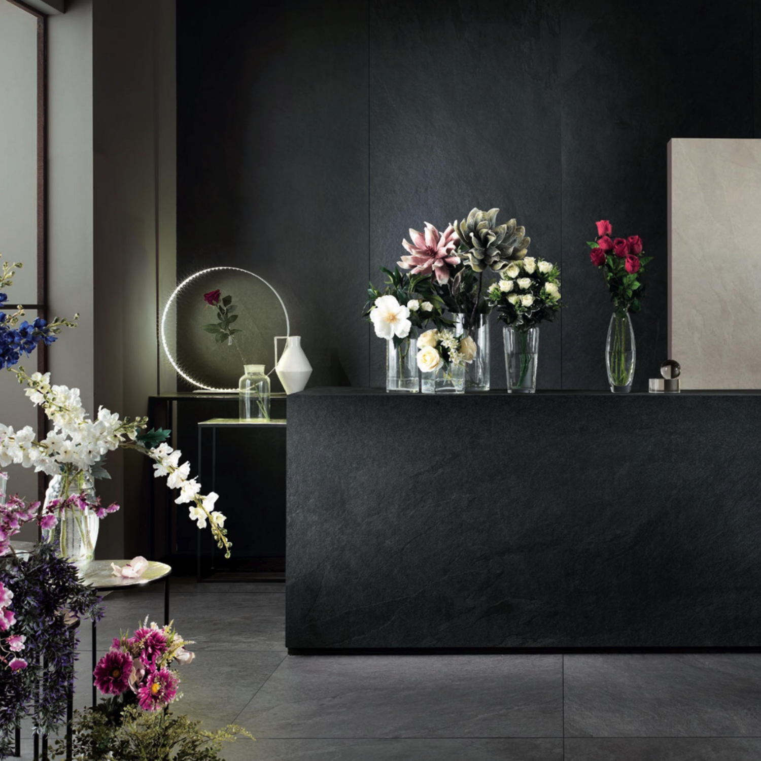 A modern room featuring the Waterfall - Dark Flow dark stone counter, clear vases with assorted flowers, a round backlit mirror, and additional floral arrangements on the floor. Minimalist decor combines with luxurious elegance for a refined atmosphere.