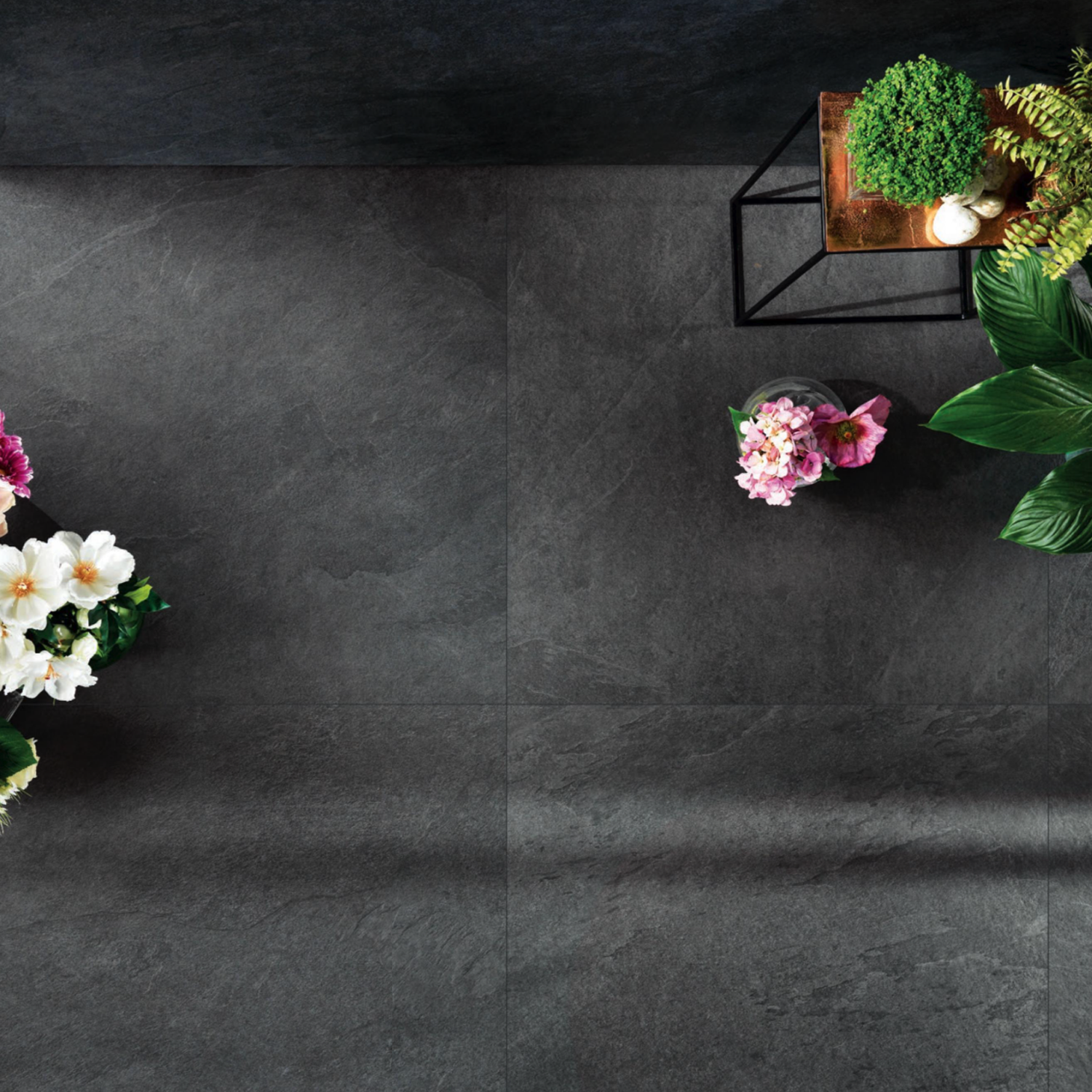Waterfall - Gray Flow tiles in dark gray are shown from above, bordered with potted green plants and pink-and-white flowers.