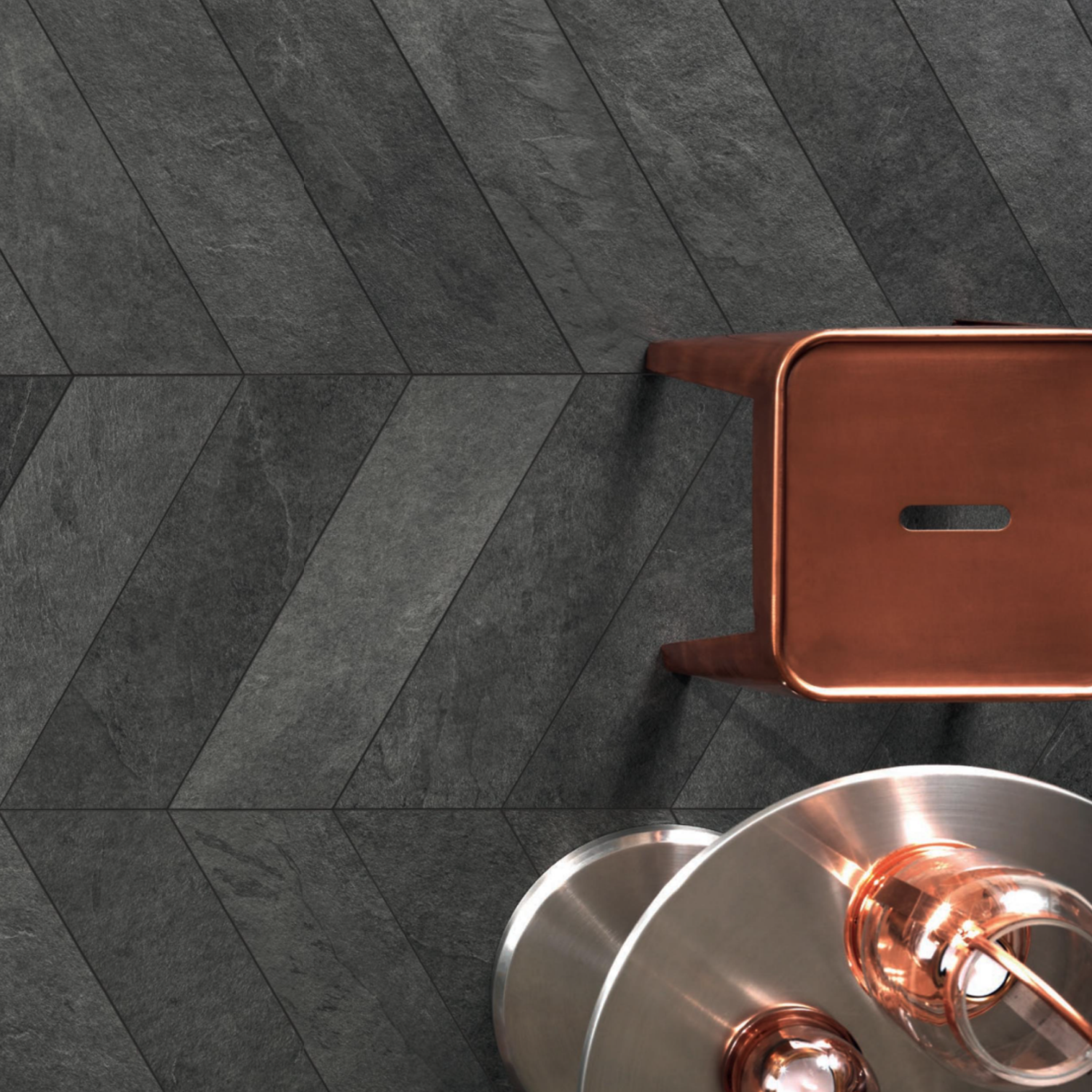 A copper stool sits next to metallic light fixtures on a floor with Waterfall - Gray Flow chevron-patterned tiles.