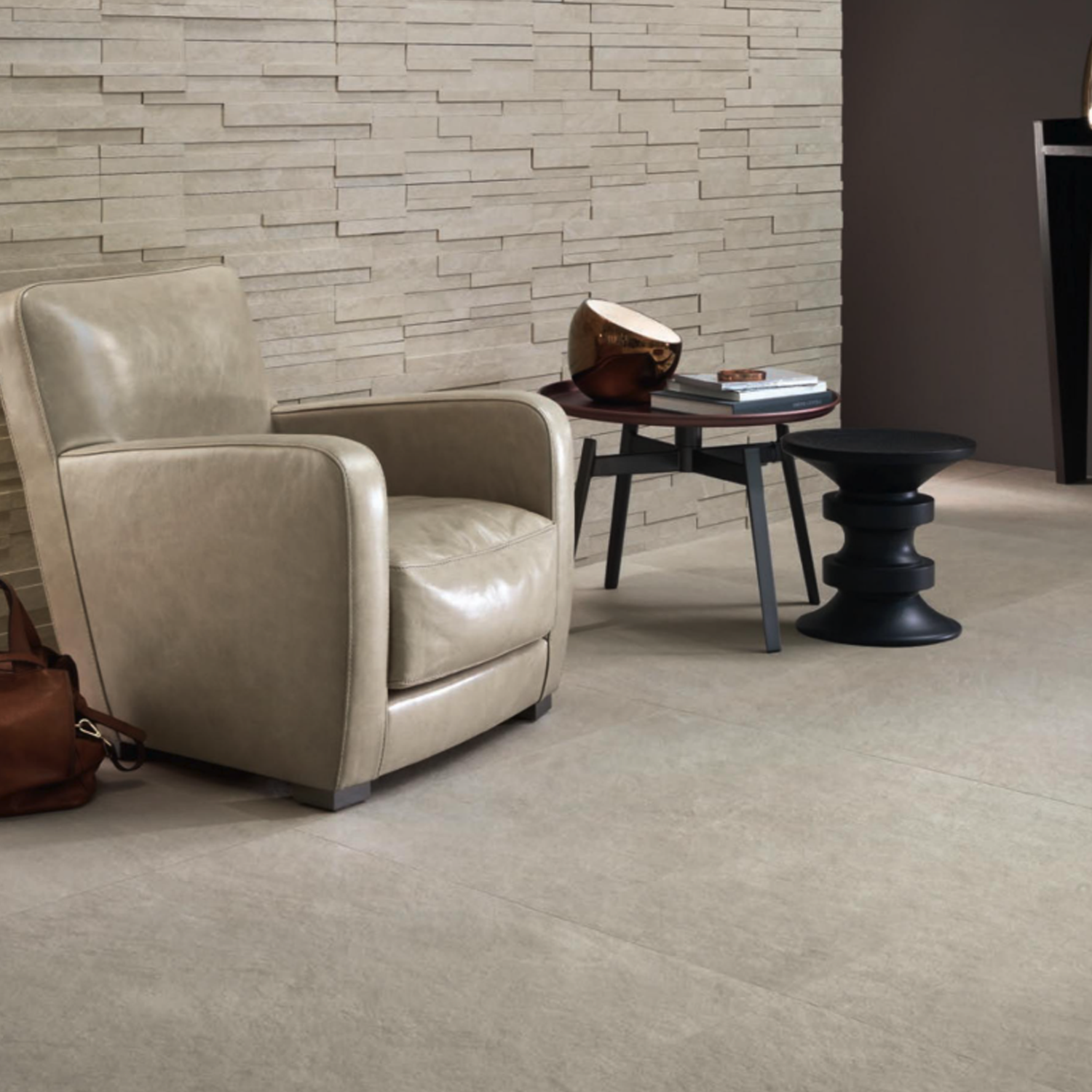 A Waterfall - Ivory Flow armchair in beige leather sits beside a small table holding books and a bag, next to a modern black stool, all arranged against a light stone accent wall in a contemporary room.