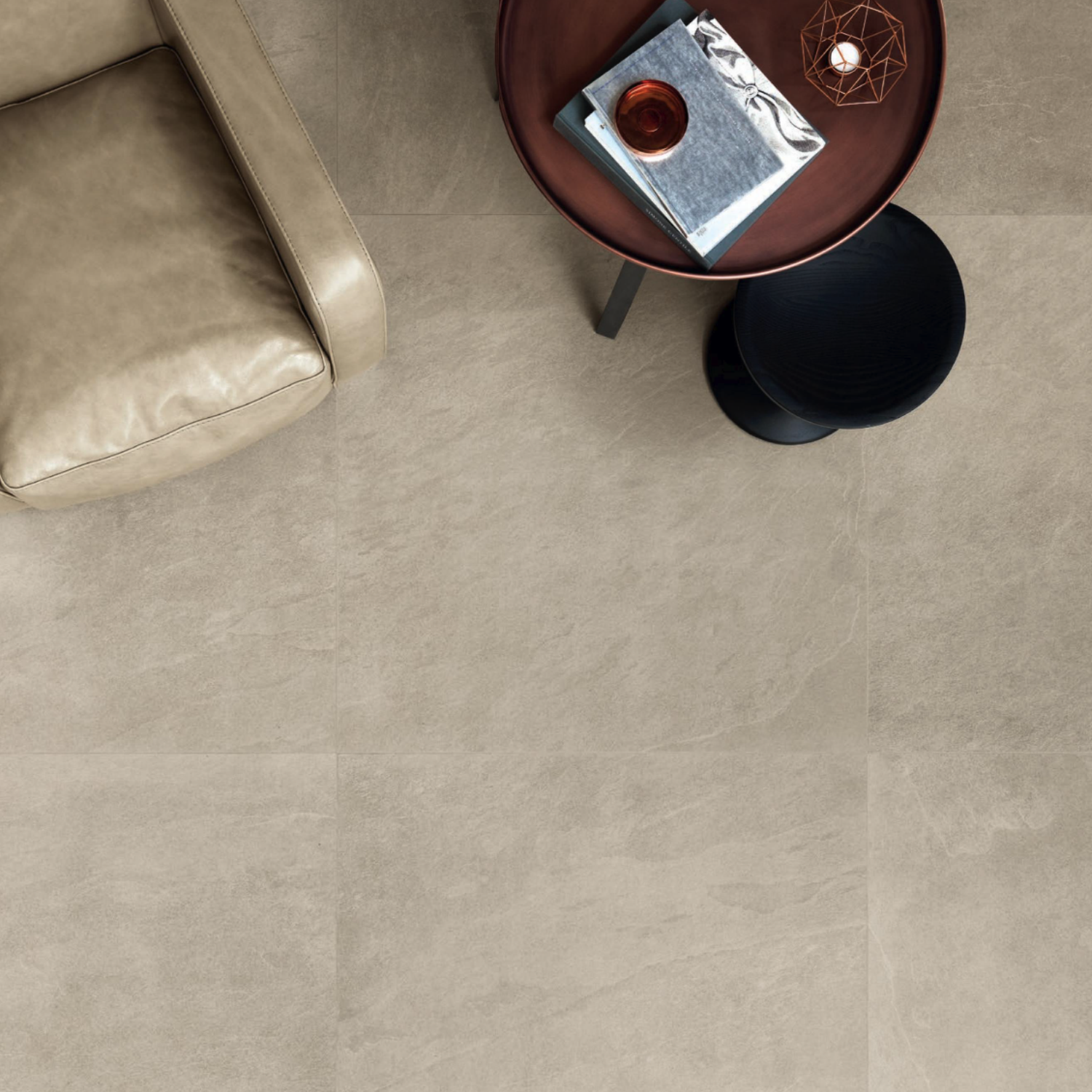 An ivory Waterfall - Ivory Flow leather chair sits beside a round wooden table topped with a blue notebook, candle, dish, and a small geometric ornament, all set on large, light stone tiles.