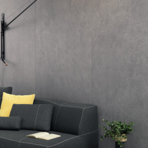 A "Waterfall - Silver Flow" wall lamp extends above a dark gray sofa with yellow and black pillows, which sits against a gray wall. An open book rests on the sofa, while a green potted plant is placed on the floor nearby.