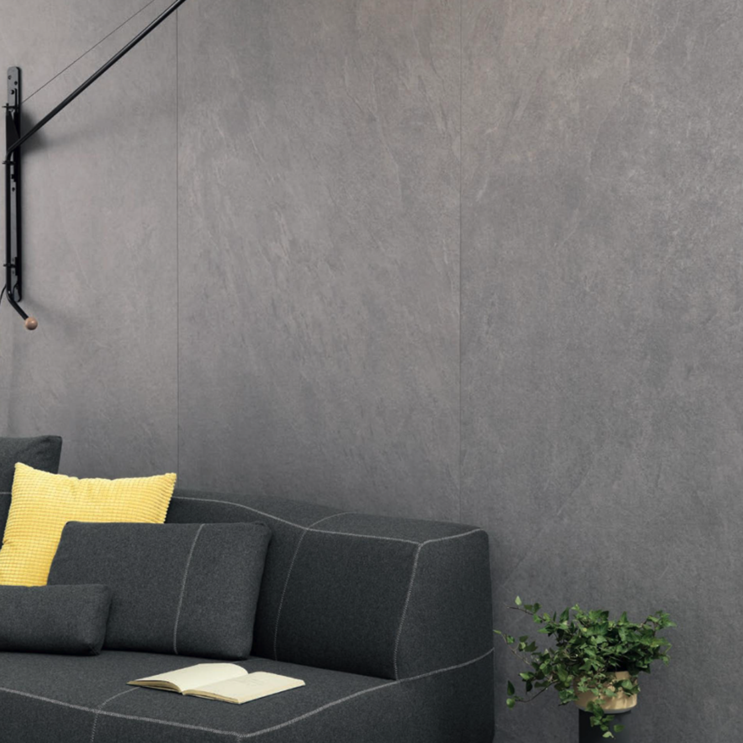 A "Waterfall - Silver Flow" wall lamp extends above a dark gray sofa with yellow and black pillows, which sits against a gray wall. An open book rests on the sofa, while a green potted plant is placed on the floor nearby.