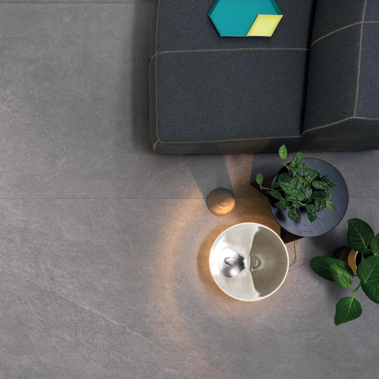 A Waterfall - Silver Flow sofa sits on a gray stone floor, accompanied by colorful books, a small potted plant on a black stand, a white bowl with a spoon, and a wooden sphere, all illuminated by soft light.