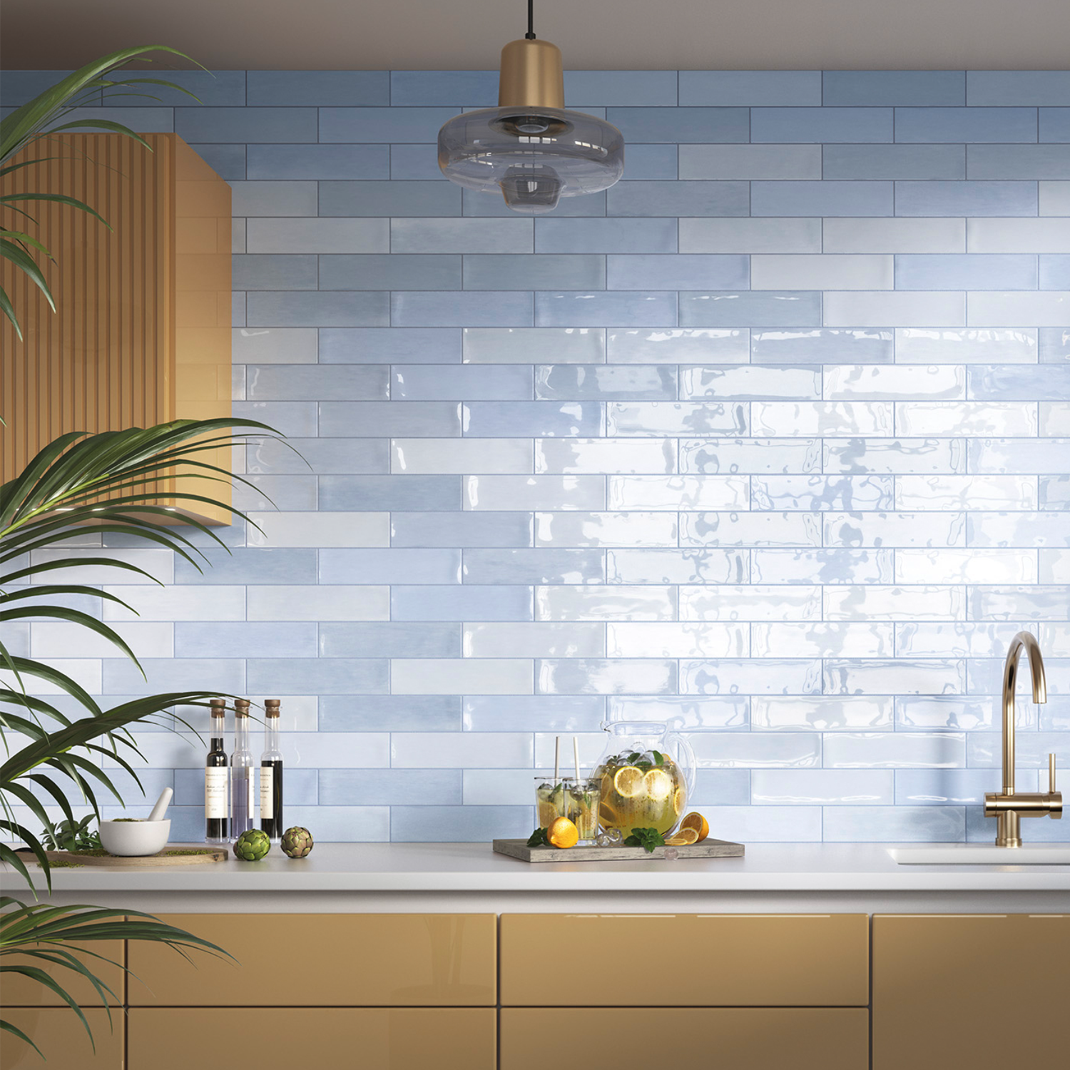 Modern kitchen featuring a Zellige - Arctic glossy light blue subway tile backsplash, gold faucet, glass pendant light, wooden cabinets, and countertop adorned with a pitcher of citrus water, limes, and bottles of oil. A green plant sits in the foreground.