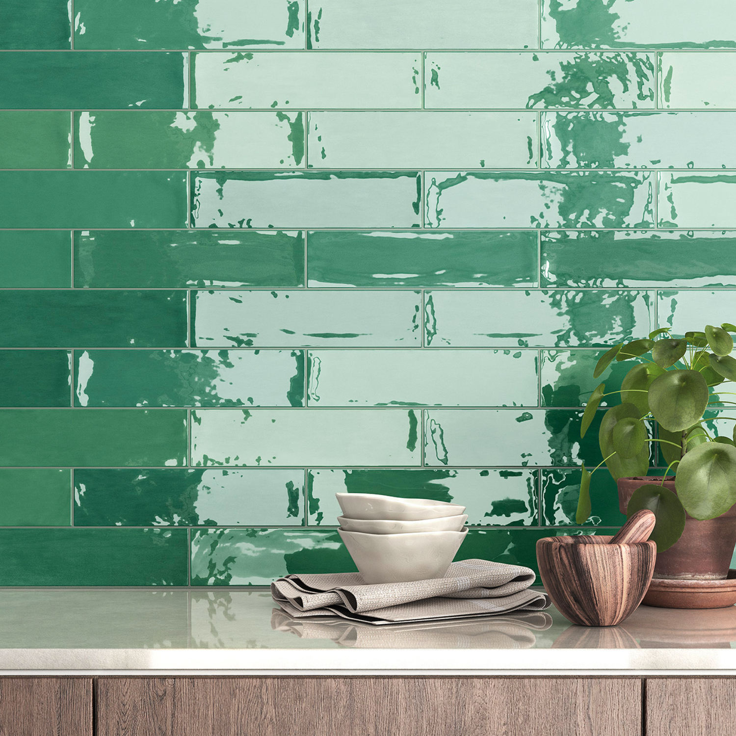 Zellige - Green subway tiles create a glossy backsplash in the kitchen, complemented by a countertop displaying stacked white bowls, a striped towel, a small wooden bowl, and a potted plant with round leaves.