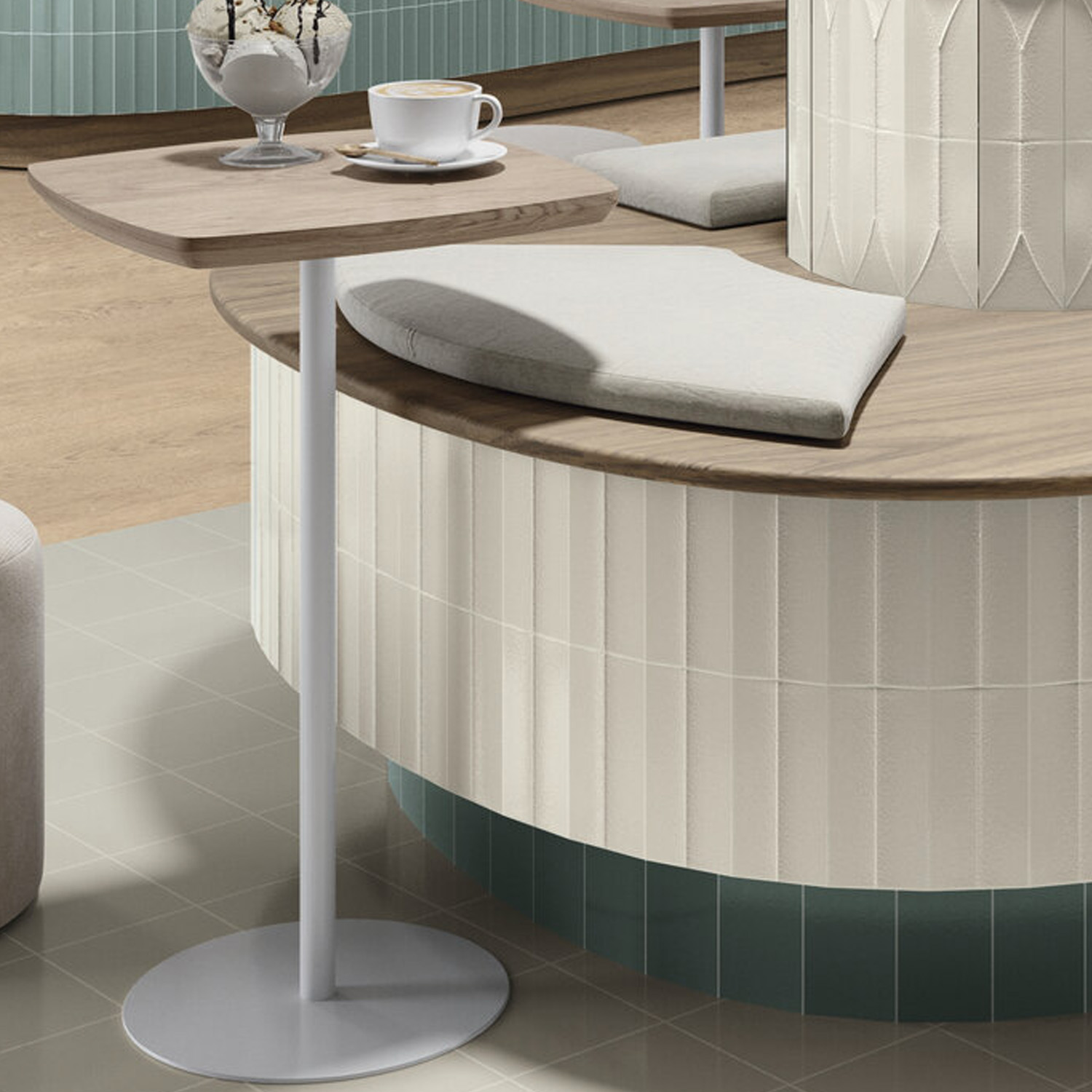 A modern cafe setting features the Arka - Arka Bone, a square wooden side table holding a cup and saucer. Beside it sits a curved bench with light cushions, all set against textured cream and teal tiled walls and light wood flooring.