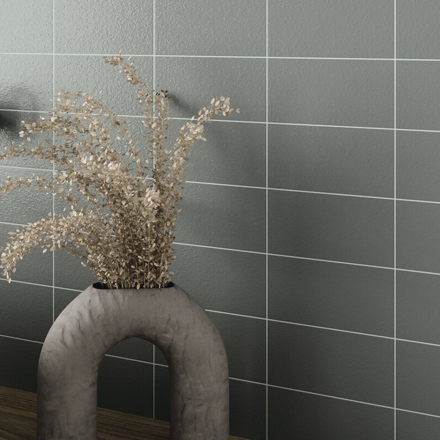 An Arka - Arka Steel vase with dried branches sits on a wooden surface in front of a wall with large, dark gray tiles and white grout lines.