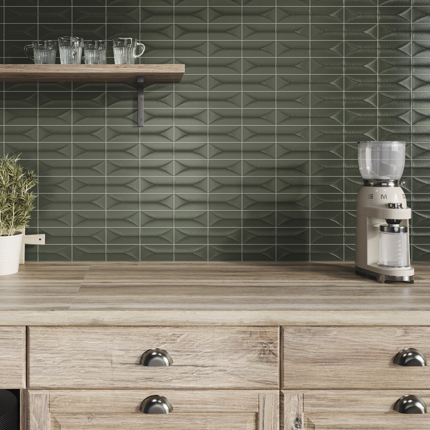 A wooden kitchen counter with drawers, a potted plant, a glass container appliance, and a floating shelf holding glassware. The backsplash features Arka - Forma Green geometric tiles.