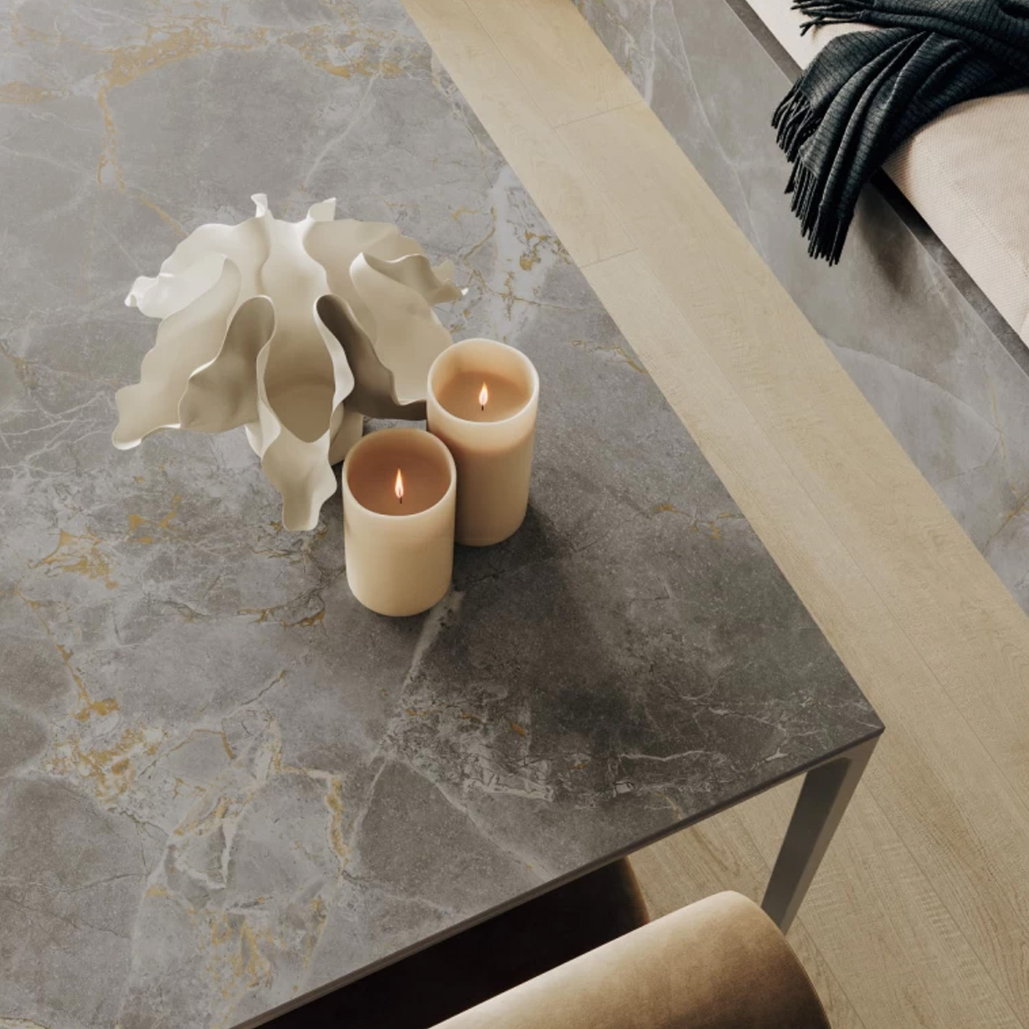 Two lit Fior Di Bosco beige candles and a white decorative object sit on a gray marble table. A beige bench and a folded dark green blanket are visible in the background.