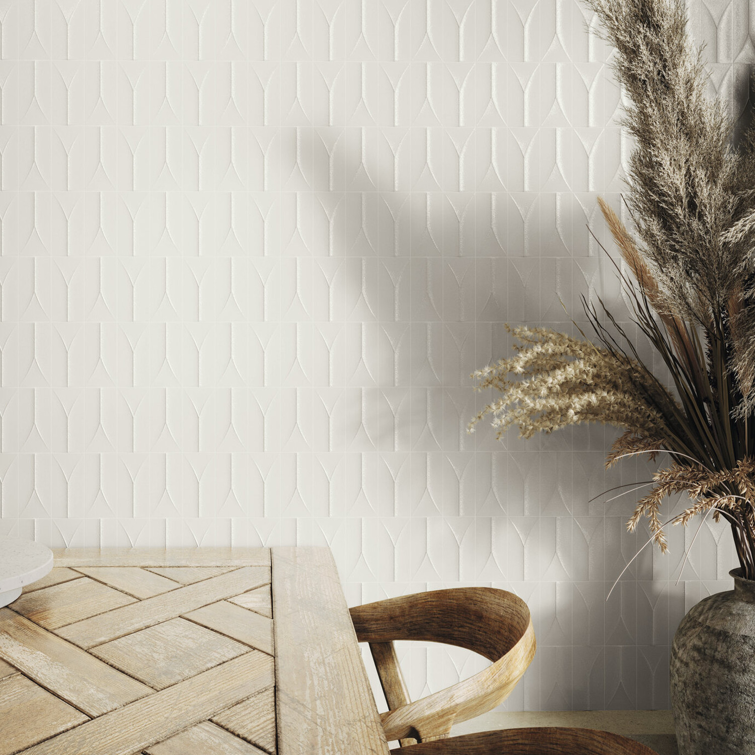 An Arka - Forma White textured wall with geometric patterns forms the backdrop for a wooden table and chair, while a vase filled with dried grasses and pampas stands on the right, casting a shadow.