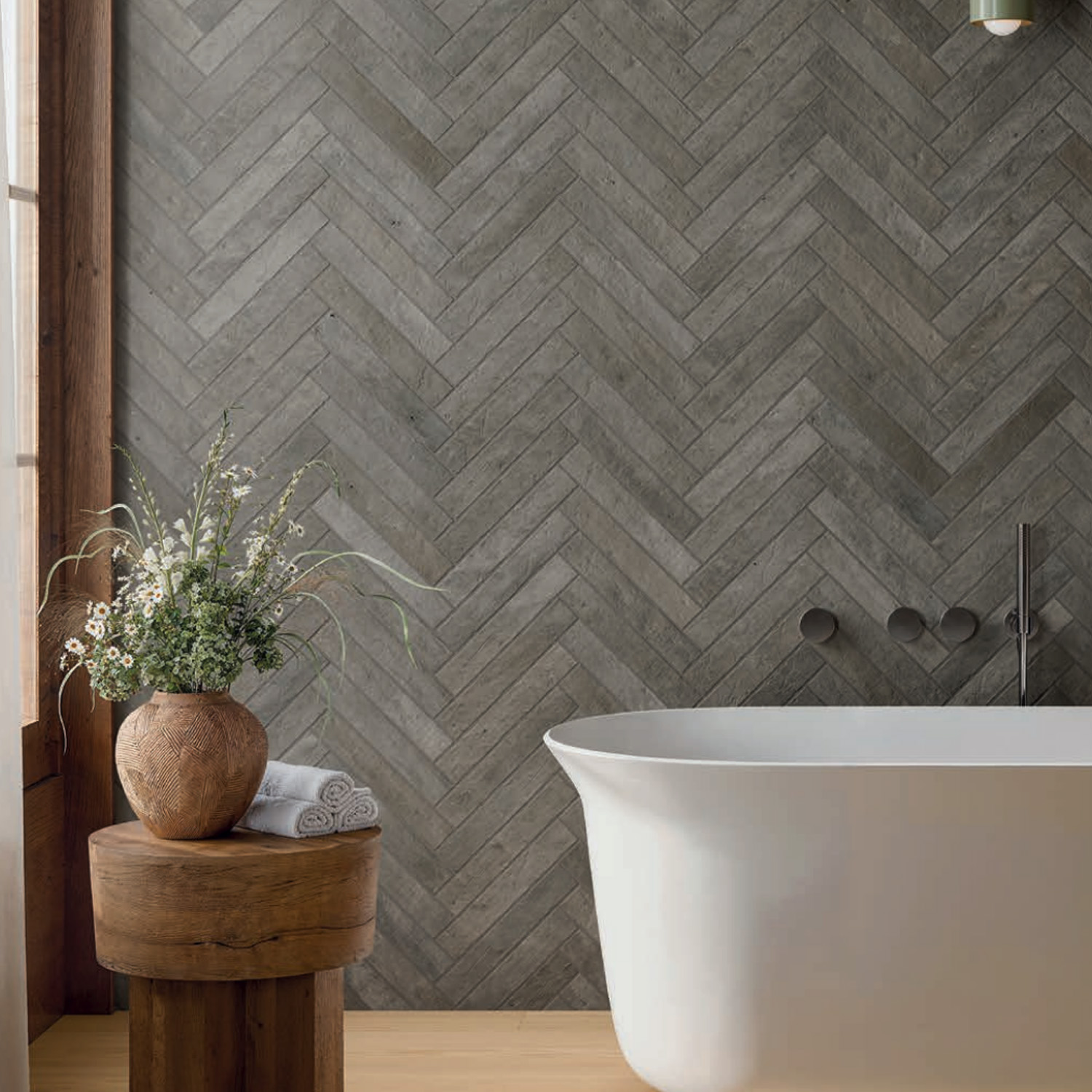 A modern bathroom features the Tyrrhenia - Lagoon white freestanding tub, accompanied by a wooden stool topped with a vase of greenery, set against a backdrop of gray herringbone tile. Natural light streams in from the left window.